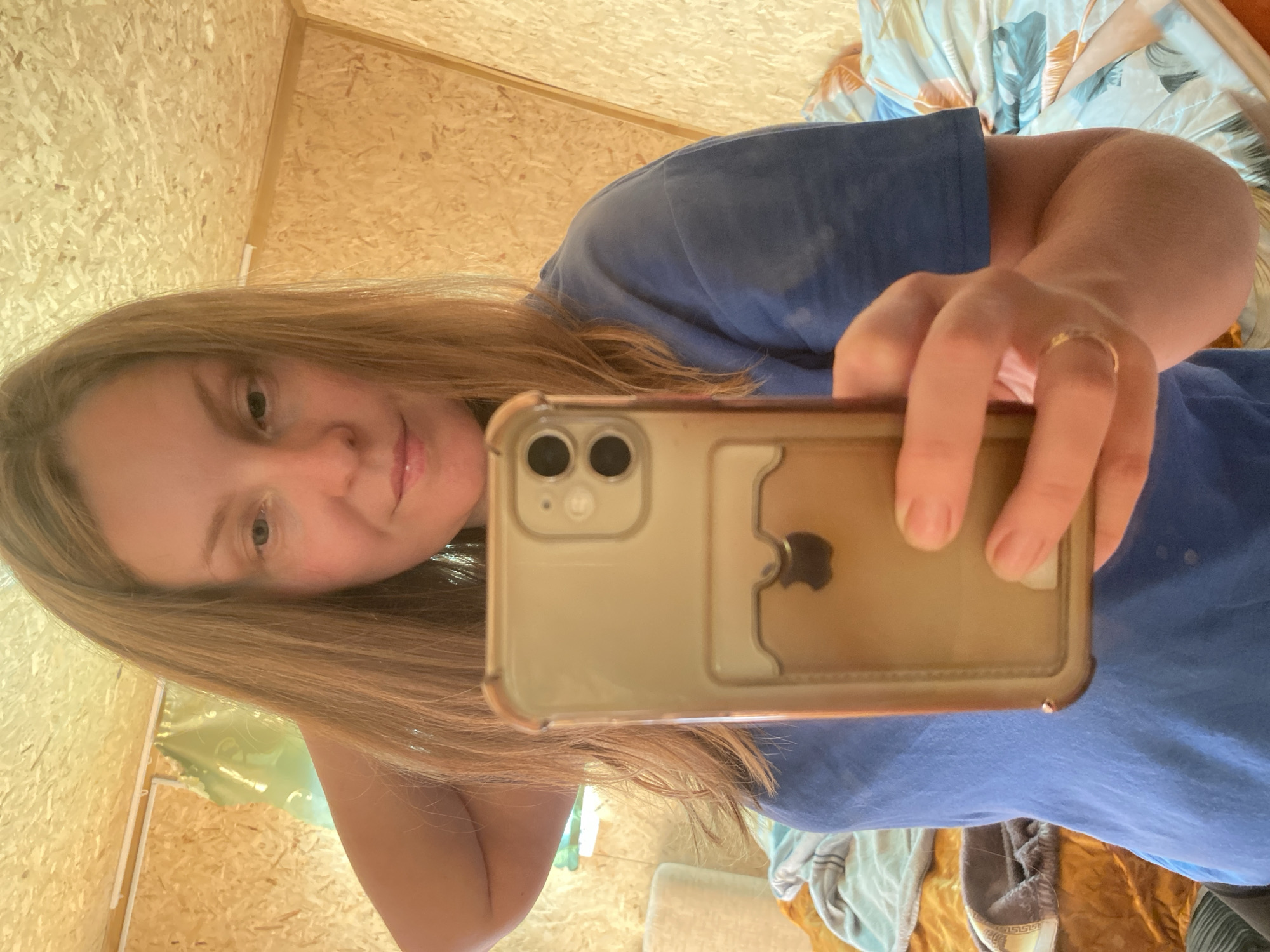 A woman is depicted taking a selfie with an iPhone in a room in the city of Chaikovsky, Russia. The woman, with long light brown hair and wearing a blue t-shirt, holds the phone in her right hand, displaying a ring on her right ring finger. The iPhone, identifiable by its dual camera system and an Apple logo visible through a discolored clear case with a card slot, occupies the lower center of the image. The room features walls and a ceiling constructed from oriented strand board (OSB) panels. In the background, a bed is visible with bedding; one section displays a pattern of blue and orange/brown foliage, while another section on the left is golden-yellow with blue fabric draped over it. A piece of light green fabric or plastic is suspended in the upper left background, adjacent to a white linear trim or cable channel. A white cylindrical object is partially visible on the left side of the bed.