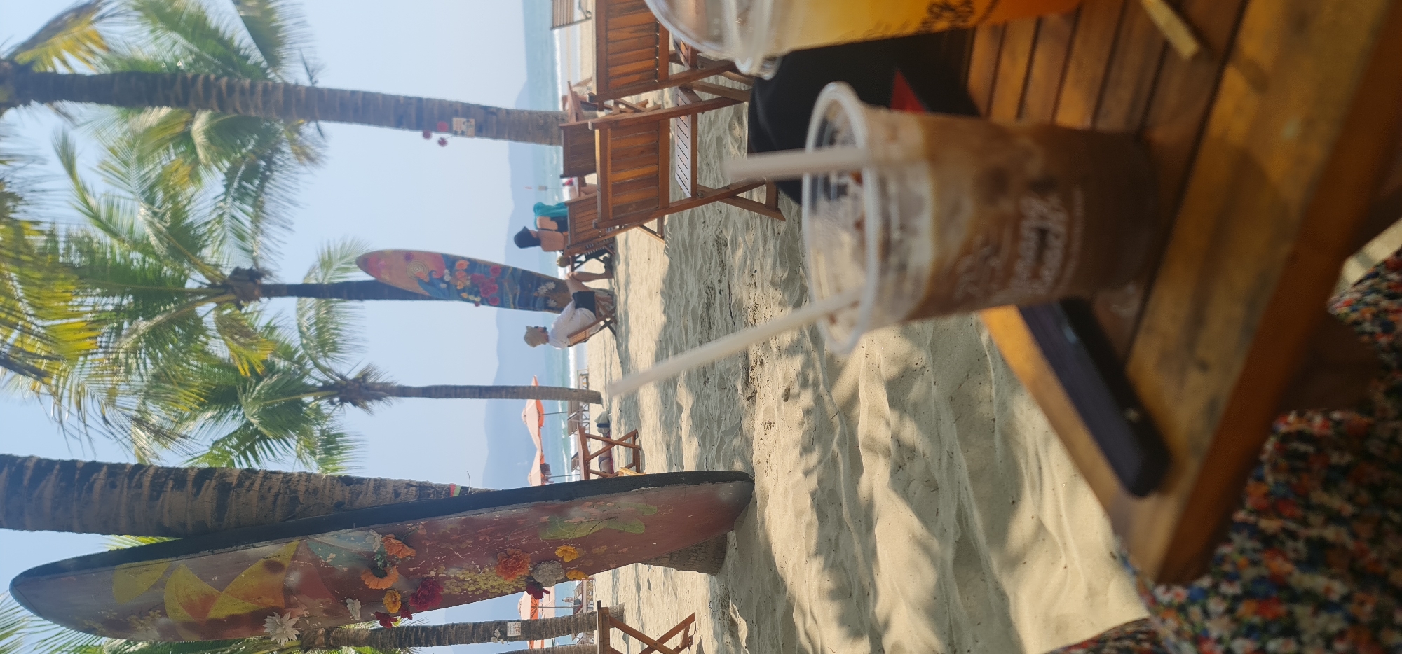 The image captures a beach scene in Đà Nẵng, Vietnam. In the foreground, a partially visible plastic cup with a straw sits on a wooden table, indicating a café or beach bar setting. A person is sitting at the table with a floral print garment visible. The sandy beach stretches out, populated with lounge chairs, beachgoers, and palm trees. Surfboards decorated with flowers are displayed against the palm tree trunks. The sea is visible in the background, with some distant mountains or landmasses in the far background. Two people are sitting on chairs on the beach. The sky is bright and clear.