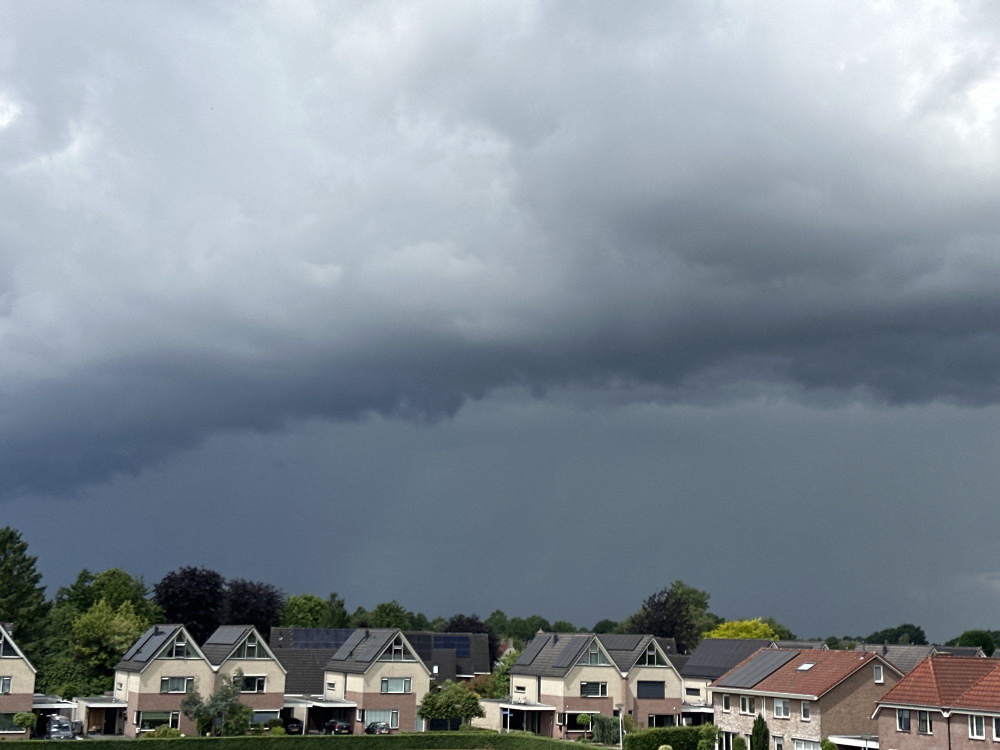 This image captures a suburban landscape under a dramatically overcast sky, indicative of an approaching storm. The upper two-thirds of the frame are dominated by heavy, dark grey storm clouds, with lighter grey clouds visible higher up and towards the left. The clouds appear thick and dense, with visible rain streaks or virga falling from the darkest, lowest part of the cloud layer on the left side, suggesting impending or ongoing precipitation.
Below the menacing sky, a row of modern houses is visible. These are primarily two-story homes with pitched roofs, many of which are equipped with numerous solar panels, highlighting a trend towards sustainable energy. The houses feature a mix of light-colored facades and brickwork. Lush green trees and other foliage are abundant, creating a verdant backdrop behind and between the houses. Some vehicles are partially visible parked near the homes.
The overall atmosphere is one of anticipation, with the dark clouds casting a somber, dramatic light over the residential area. It is clearly daytime, but the heavy cloud cover significantly darkens the scene. The architecture and dense suburban setting are consistent with the provided location context of Almelo, Netherlands. There is no visible text in the image.