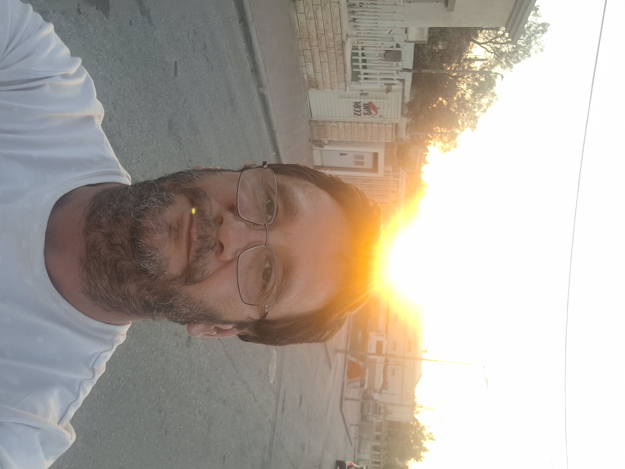 A selfie captures a man with glasses and a beard against a backdrop of a street in Yerocham, Israel, during sunset. The sun is low in the sky, casting a bright orange glow that washes out much of the background. In the foreground, the man wears a white t-shirt. Behind him, a residential street is visible, with houses and fences lining the road. A street lamp stands on the right side of the image. The overall scene is indicative of a warm evening in a quiet urban environment.