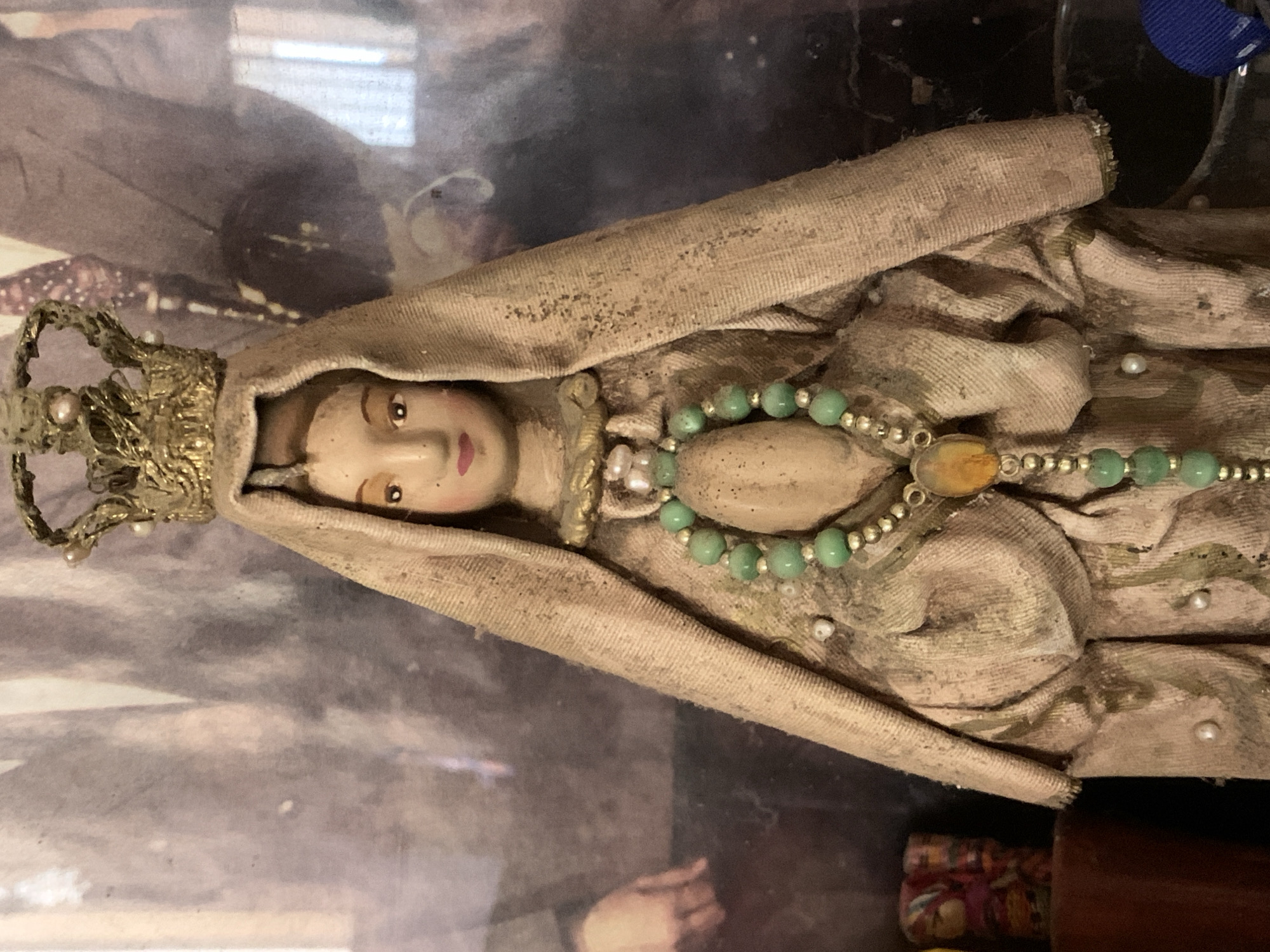 This media file displays a religious figurine, likely depicting the Virgin Mary, positioned horizontally within the frame. The figurine features a pale, doll-like face with neatly painted brown eyes, eyebrows, and pink lips. It is adorned with an ornate, gold-colored crown, intricately detailed and studded with small pearls.
The figure is draped in an aged, light beige or light brown fabric, which appears worn and possibly dusty, showing some discoloration and dark speckles. Around its neck and chest area, a necklace or rosary is visible, composed of light green spherical beads interspersed with smaller silver-colored beads, and a central oval-shaped, amber-colored stone. Several small white pearls are also scattered on the fabric drapery.
The background is blurred and indistinct, suggesting an indoor setting. It appears to be a reflection or a framed image, with shadowy areas and some lighter patches; one section vaguely resembles a human hand. The overall impression is that of an old, revered artifact, possibly displayed in an altar or case. No activity is taking place, and no visible text is present. This object is located in Santa Ana, Venezuela.