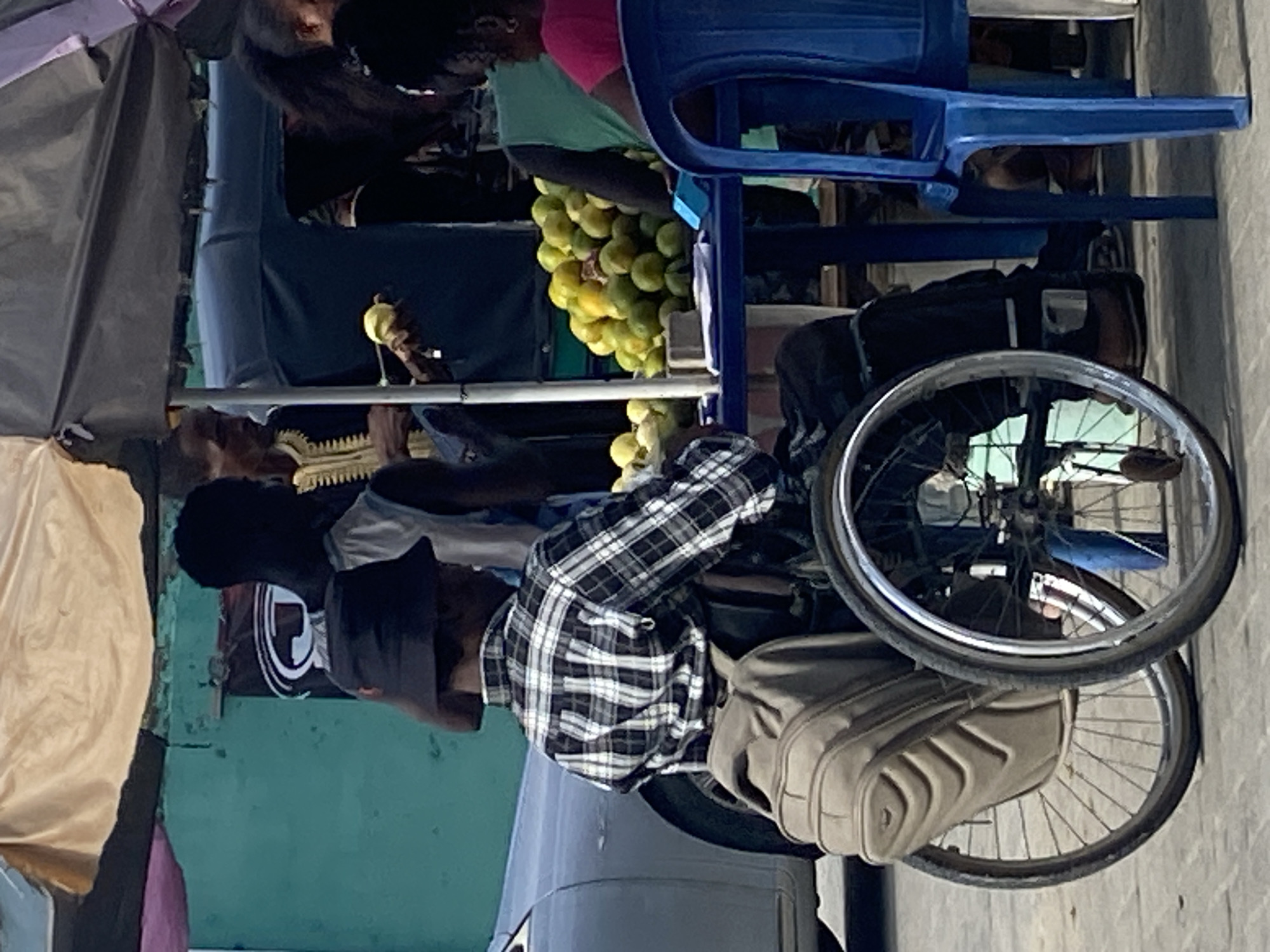 In Ado, Nigeria, a man in a wheelchair, wearing a plaid shirt and dark hat, faces right. He appears to be selling limes from a display rack attached to the wheelchair, with another person holding a lime next to him. There is a blue plastic chair next to the display. The background includes a building facade, a parasol, and parts of other people. The scene occurs on a sidewalk.