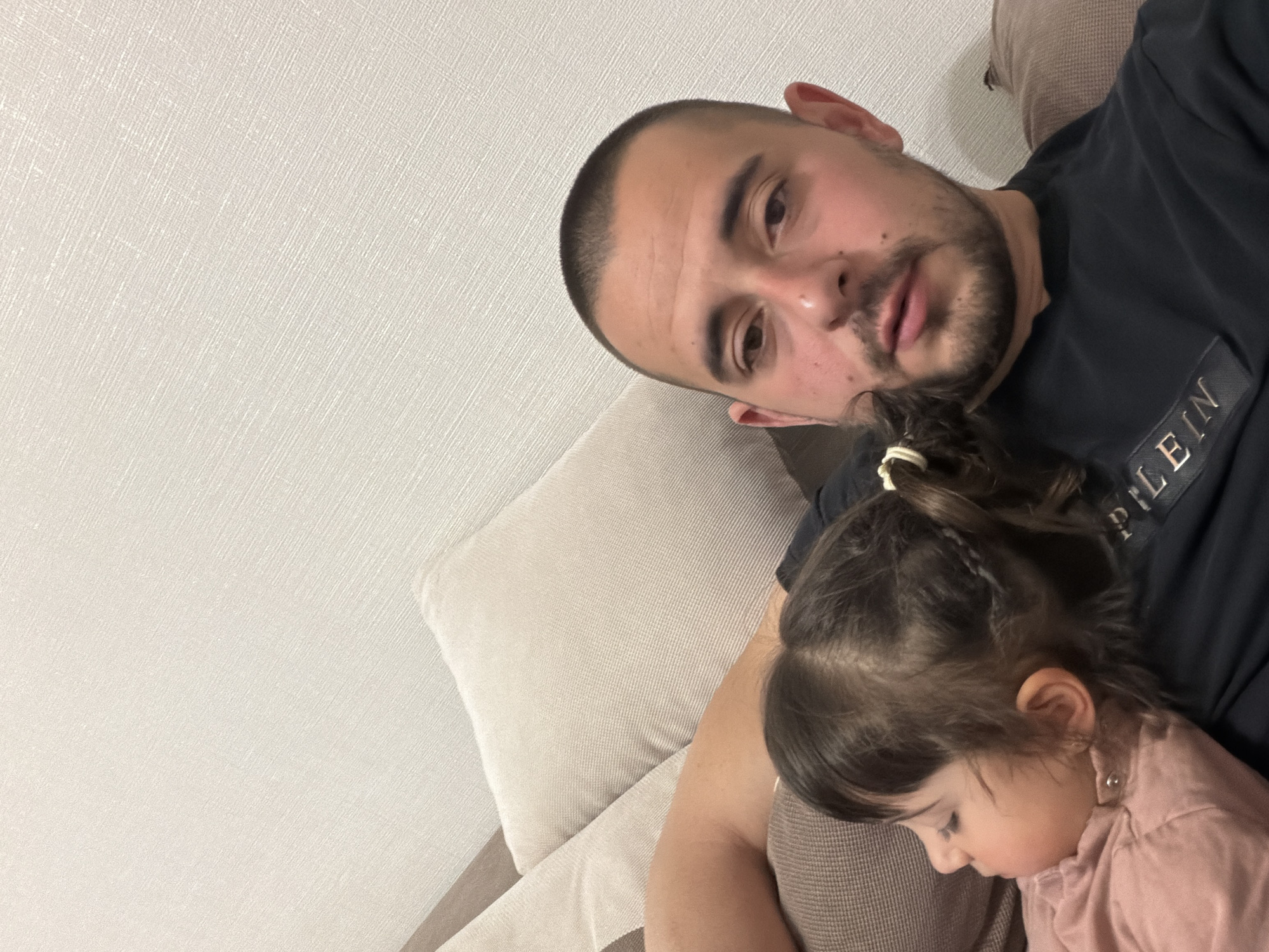 An intimate indoor portrait captures a man and a young child resting together on a sofa. The image is taken from a slightly high and tilted angle, looking down at the subjects.
The man, positioned on the right side of the frame, has short, buzzed dark hair and a dark beard or stubble. He is wearing a black t-shirt, on which a partial word "PLEIN" is visible, likely part of a brand name. He looks directly towards the viewer with a calm, neutral expression. His arm is visible, supporting the child.
Resting on the man's chest or shoulder, mostly in the lower right quadrant, is a young girl with dark hair tied in a small ponytail. She appears to be sleeping or resting peacefully, with her eyes closed and her face turned away from the camera, nestled against the man. She is wearing a light pink top.
The setting appears to be a cozy indoor space, possibly a living room. A light-colored, textured wall fills the left and top portions of the background. A beige or light brown sofa with cushions is visible, providing the resting place for the subjects. The lighting is soft and even, suggesting an indoor environment without strong natural light. The overall mood is one of quiet affection and tranquility.