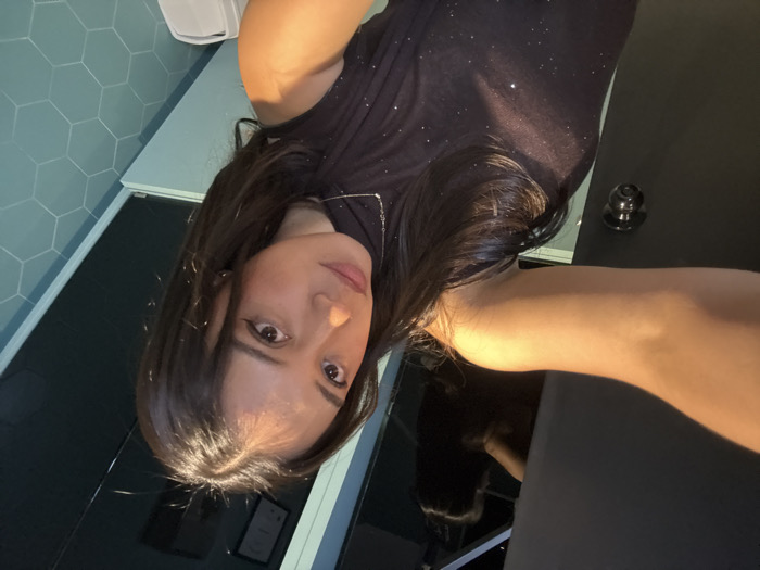 A young adult female is depicted in a close-up selfie taken indoors. She has dark, shoulder-length hair with a middle part and is wearing a dark, sleeveless top with subtle glitter. A thin silver-colored necklace is visible. Her right arm is extended into the foreground, holding the camera, with the hand out of frame. She looks directly at the camera with a neutral expression.
The background on the left features a dark, solid-colored door or partition with a dark metallic doorknob. Above this, a reflection shows the top of a head, likely the subject's, and an overhead light fixture. The background on the right consists of a wall with light blue hexagonal tiles, above which is a dark, possibly reflective, surface with a framed certificate visible. A white object, possibly a trash bin or toilet, is partially visible at the bottom right. The specific location within San Bernardino Tlaxcalancingo, Mexico, cannot be determined from the visual information provided in the image.