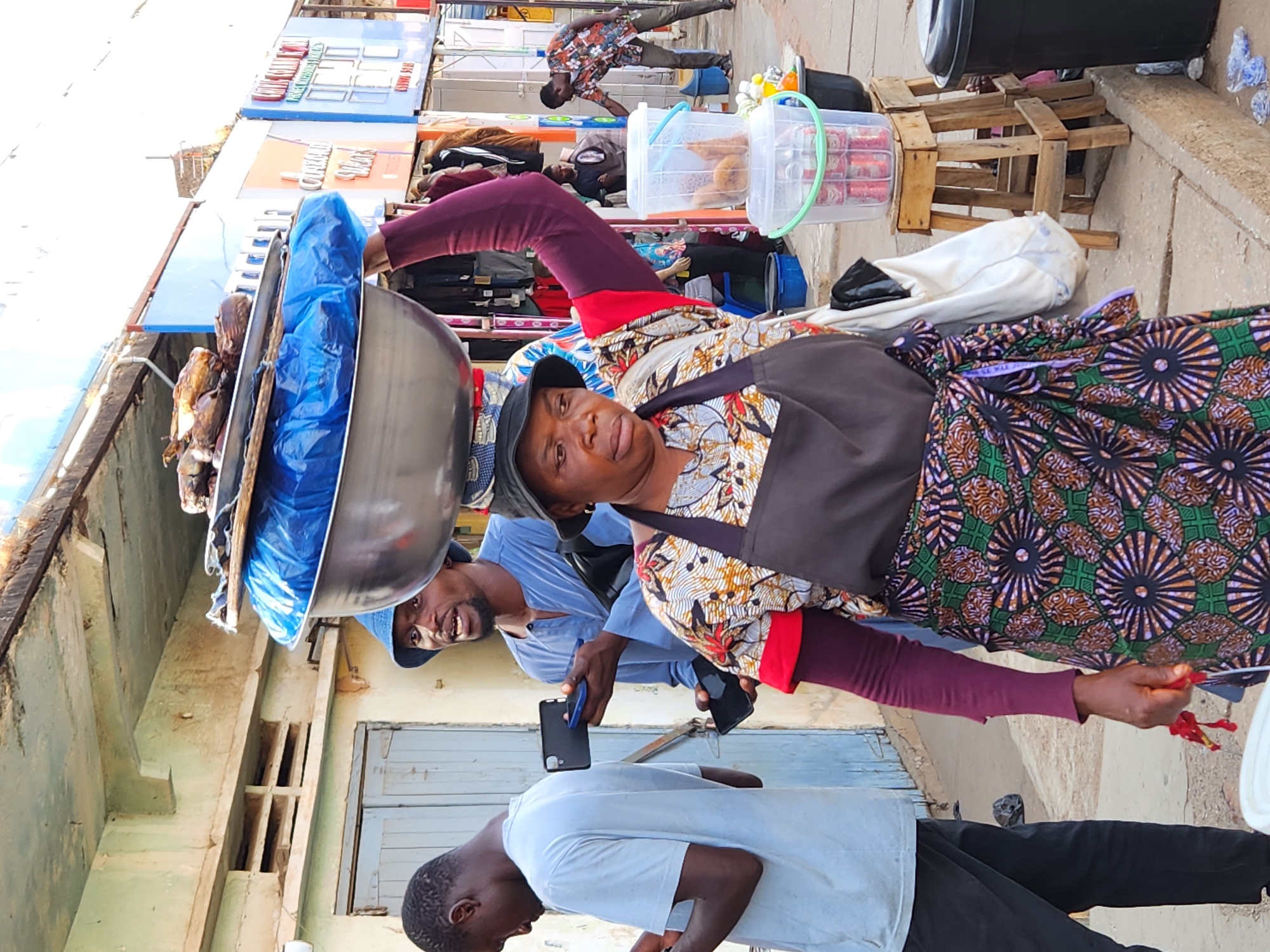 The image depicts a scene in Jos, Nigeria. A woman wearing a patterned dress, a dark apron, and a hat is carrying a large metal bowl on her head that contains food, possibly fish, covered with a blue plastic sheet. She is the primary focus and is surrounded by people and objects typical of a street market.
To the right, there are stacked wooden pallets, containers, and cans. A man in a blue shirt is visible behind her, holding a cell phone and looking in her direction. Another man, wearing a light-colored shirt and dark pants, is visible in the lower part of the image.
The background includes buildings with closed doors. Overhead, there are signs and awnings indicating stalls or shops. The image orientation is rotated, but objects and details allow for identification of the scene.