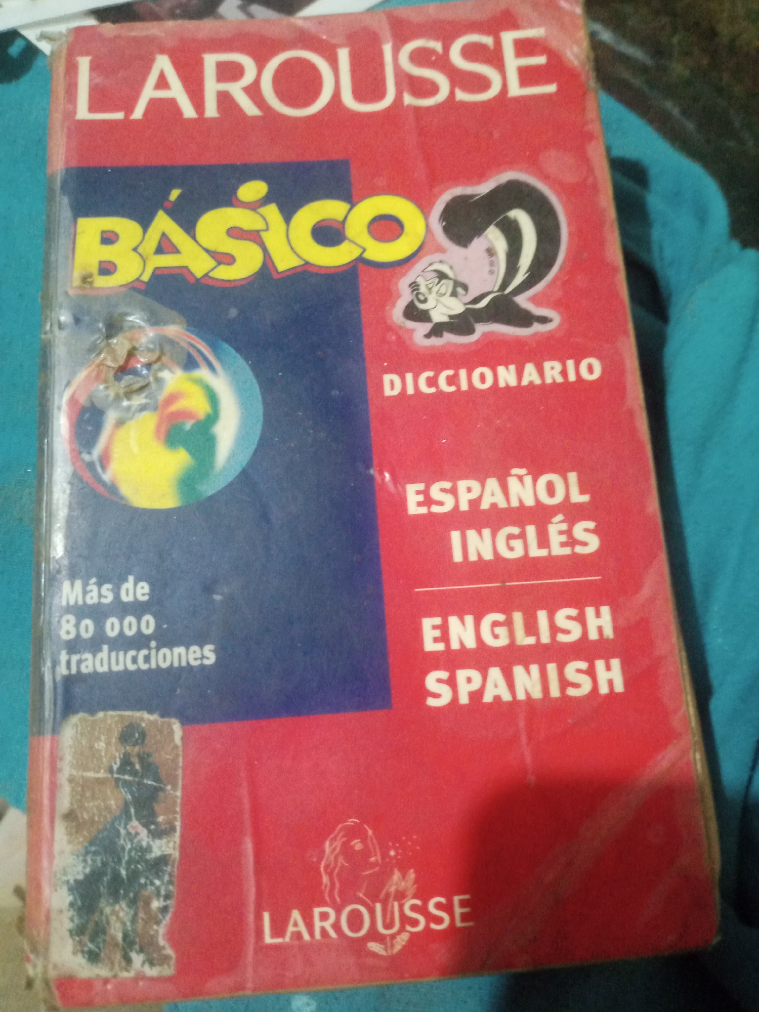 The image shows a red and blue book titled "LAROUSSE BÁSICO DICCIONARIO ESPAÑOL INGLÉS ENGLISH SPANISH". The book appears to be worn and possibly old. There is a cartoon skunk character on the cover. The book is placed on a blue fabric surface. The image was taken in Santa Ana, Venezuela.