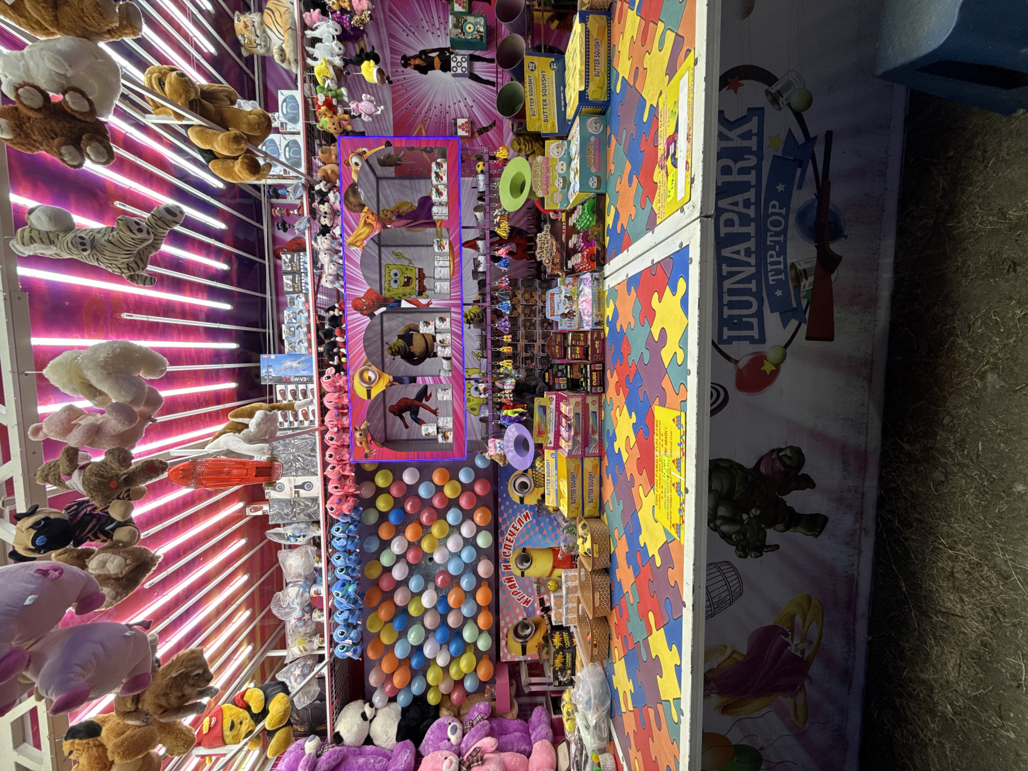 This image depicts a carnival game stall, likely at a funfair or amusement park. The stall is brightly lit with pink neon lights and features a variety of stuffed animals hanging from above, including plush dogs, tigers, and other characters. The foreground shows a colorful, puzzle-piece-like surface, a common feature of such attractions.
Behind this surface, there's a game area with shelves stocked with various prizes, including boxes of "BUTTER SQUISHY" toys and other small items. There's also a backdrop with cartoon characters like SpongeBob SquarePants, Shrek, and Rapunzel, suggesting a family-friendly theme. A target shooting game, indicated by a rifle image on the stall's facade and cans, is also present.
The stall's signage reads "LUNAPARK TIP-TOP". The overall atmosphere is one of entertainment and leisure, typical of a carnival setting. While the image doesn't explicitly state the location within Lozenets, Bulgaria, it strongly suggests it is a public amusement area within the city.