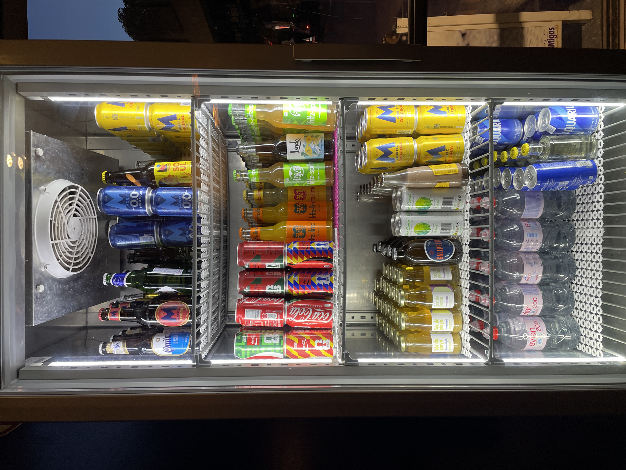 The image shows the interior of a refrigerated display case filled with various beverages, primarily located in the city of Unknown, Unknown. The display case is brightly lit from the inside.
The top shelf features several tall, blue cans stacked vertically, with some yellow and blue cans visible behind them. To the left, there are multiple bottles of beer, including some with blue and red labels.
The second shelf contains a variety of canned drinks. There are green cans, red cans with Coca-Cola branding, and some yellow and blue patterned cans. To the right, there are orange-colored canned drinks labeled "Fritz-Limo."
The third shelf holds an assortment of bottled beverages. There are numerous small, clear bottles filled with a yellow liquid, some labeled "Soc de pinyo." A few brown glass bottles and a bottle labeled "GALI PETE" are also present.
The fourth shelf contains several large bottles of Evian water, identifiable by their clear plastic bottles and pink labels.
The bottom shelf is sparsely populated with blue cans, some appearing to be sparkling water based on the "Aquarius" branding. There is also a tall, clear bottle with a yellow liquid.
The exterior of the display case is dark, with the right side of the image showing a glimpse of a dark outdoor environment, possibly a street scene at dusk or night, with some indistinct lighting. A portion of a sign reading "MIGAS" is visible on the far right.