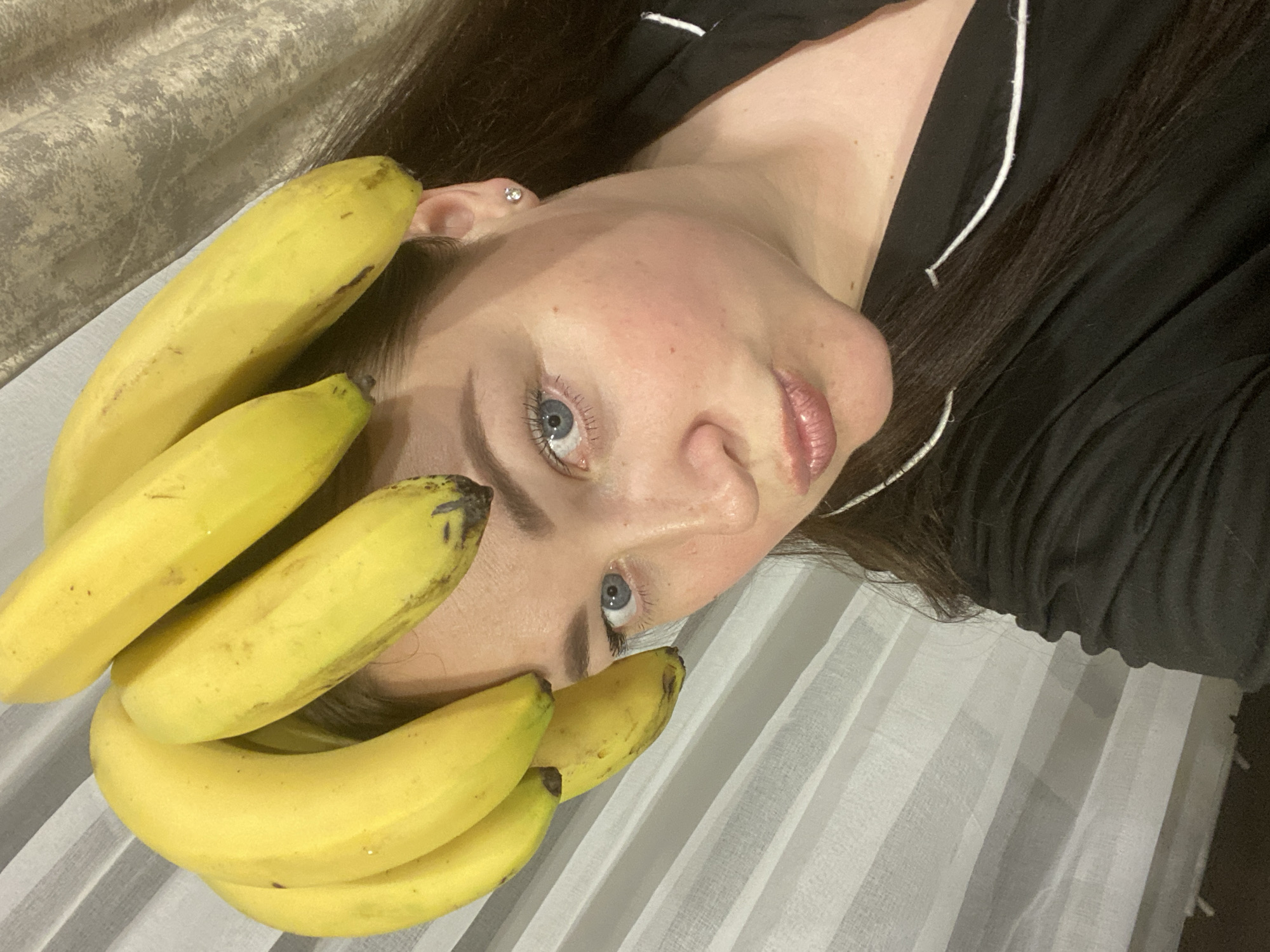 The image displays a close-up, rotated view of a person lying down with several bananas arranged around their head and face. The subject is a person with fair skin, light blue eyes, and long dark hair, wearing a dark top with white piping, possibly pajamas. A small, shiny stud earring is visible in their right ear. Their lips have a glossy appearance.
A cluster of ripe yellow bananas, some showing small brown spots, frames the person's face, with one banana resting near their ear and others positioned around their forehead and cheek. The person's eyes are open and directed towards the camera, conveying a neutral to slightly playful expression.
The background consists of two distinct fabric textures. To the left and upper part of the image, there is a patterned, light greyish-brown fabric, possibly a blanket or headboard. The right and lower part features a lighter, striped fabric, likely white or light grey, suggesting bedding or curtains. The overall scene is indoors. No specific time of day or weather conditions are discernible from the indoor lighting. There is no visible text in the image. The location context provided is Sochi, Russia, though no specific visual cues within the image confirm this.