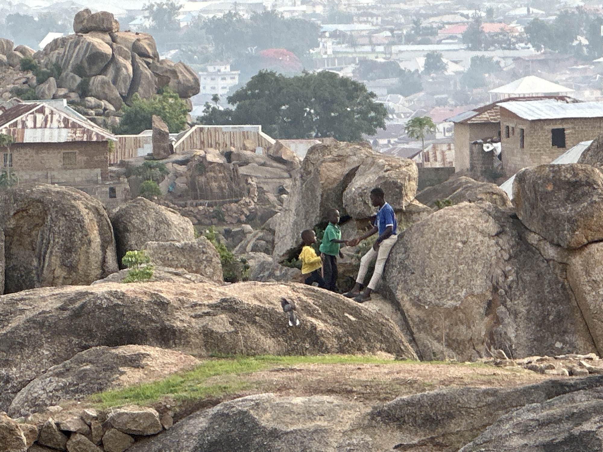 In the city of Jos, Nigeria, three young boys are interacting on a rocky landscape. One boy, wearing a yellow shirt, kneels on a large boulder. Another boy, wearing a green shirt, stands next to him. The third boy, wearing a blue shirt and light-colored pants, sits on another large boulder, appearing to shake hands with the boy in the green shirt. A small bird is perched on the same boulder as the kneeling boy. In the background, there are several buildings with corrugated metal roofs and other structures, interspersed among more rock formations and trees. The cityscape fades into a hazy distance. The foreground is dominated by textured rocks and sparse vegetation.