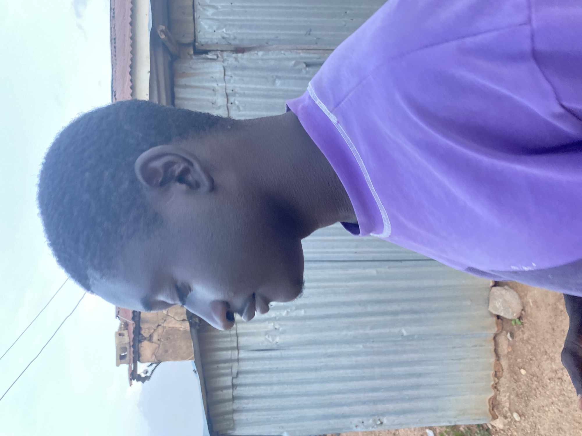 A side-profile view of a person with dark skin and short, dark hair. The individual is wearing a purple t-shirt with a white trim around the collar. The background features corrugated metal walls. Outside of a mud house in the city of Bukuru, Nigeria. The individual is facing toward the right side of the image.