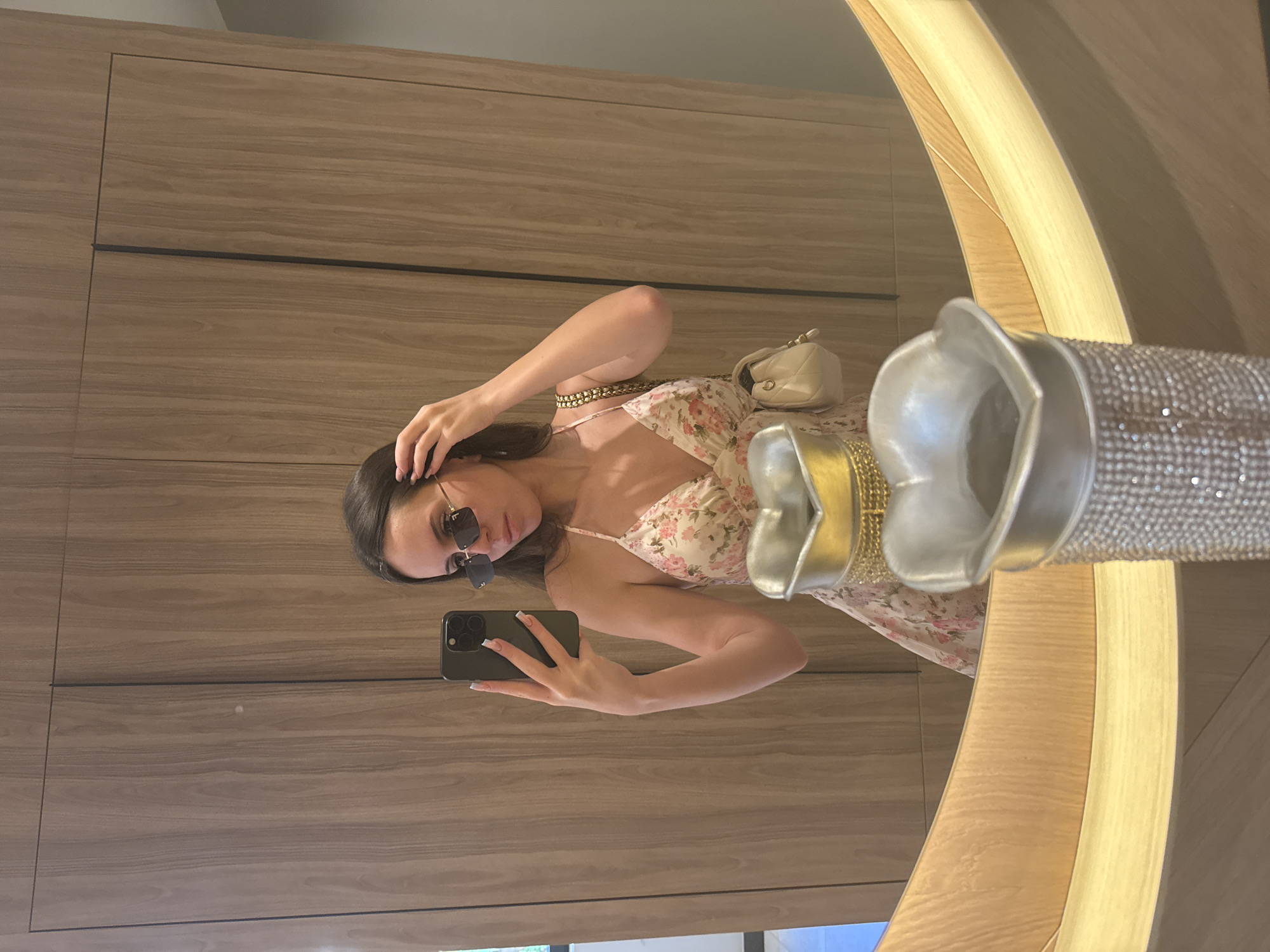 A young woman with long, dark hair is reflected in a mirror, taking a selfie. She is wearing a floral print sundress and sunglasses, and holding her hair with one hand. A small beige purse with a gold chain strap is visible on her shoulder. The mirror has a curved, wooden frame and is positioned in front of a wooden paneled wall. In the foreground, part of a decorative silver vase with a crystal-like embellishment is visible. The setting appears to be an indoor space, possibly a dressing room or hallway, in Unknown, Unknown.