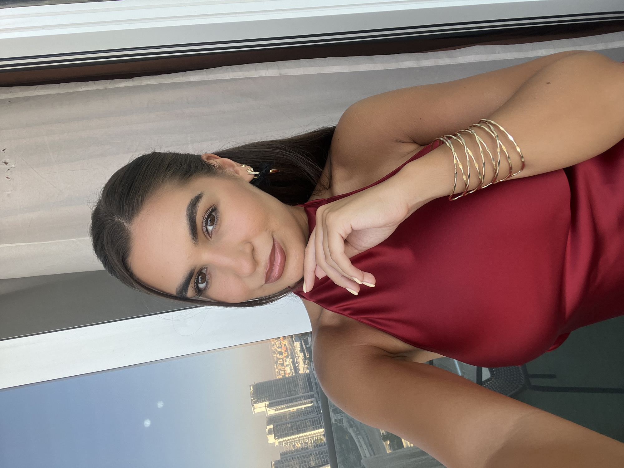 A woman is facing the camera in what appears to be a selfie. She is wearing a sleeveless red dress, with several gold bangles on her left upper arm. Her right hand is placed delicately near her neck. She has dark hair and light brown eyes.
The background reveals a cityscape, which, based on the architecture and urban layout, appears to be Dubai, United Arab Emirates. High-rise buildings dominate the view. The sky above the city is illuminated with the golden hue of what looks like either sunrise or sunset. She is in front of a window, with the window frame visible at the top of the shot.