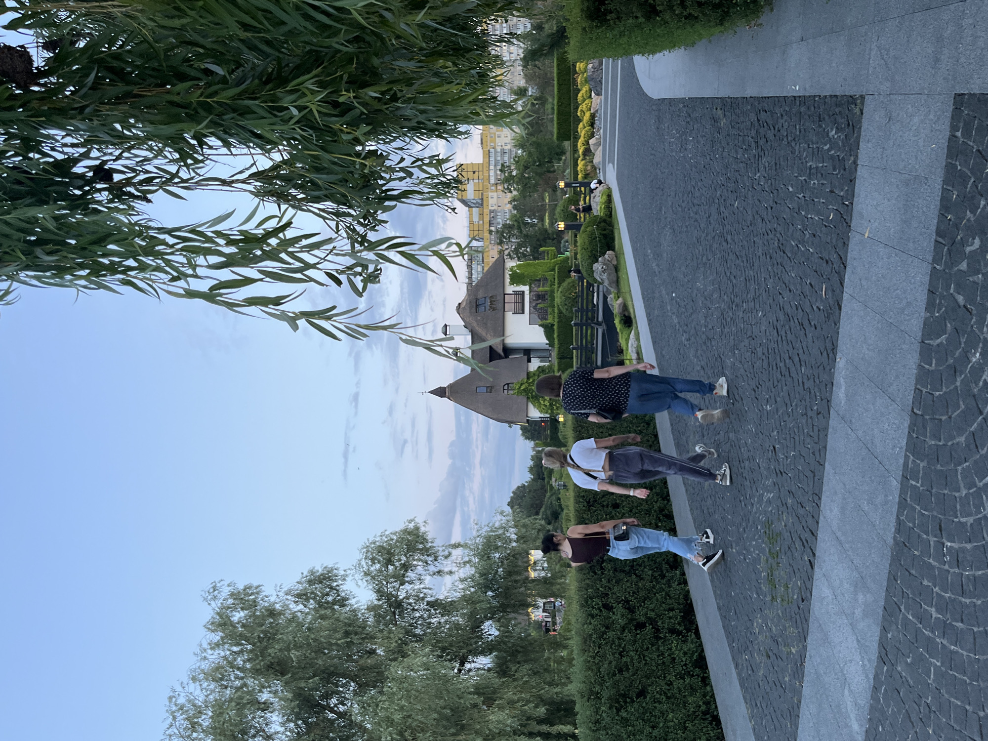 Three women walk away from the camera on a cobblestone path in a park-like setting. The woman on the left wears a dark tank top and light-wash jeans. The middle woman wears a white t-shirt and grey sweatpants. The woman on the right wears a black and white polka dot shirt and blue jeans. They are walking towards a building with a thatched roof and a modern, yellow building in the distance. The park features manicured hedges, trees, and decorative rocks. The sky is overcast and the light suggests it is either early morning or late evening. This scene is located in the park along the Dnipro River in the city of Dnipro, Ukraine.