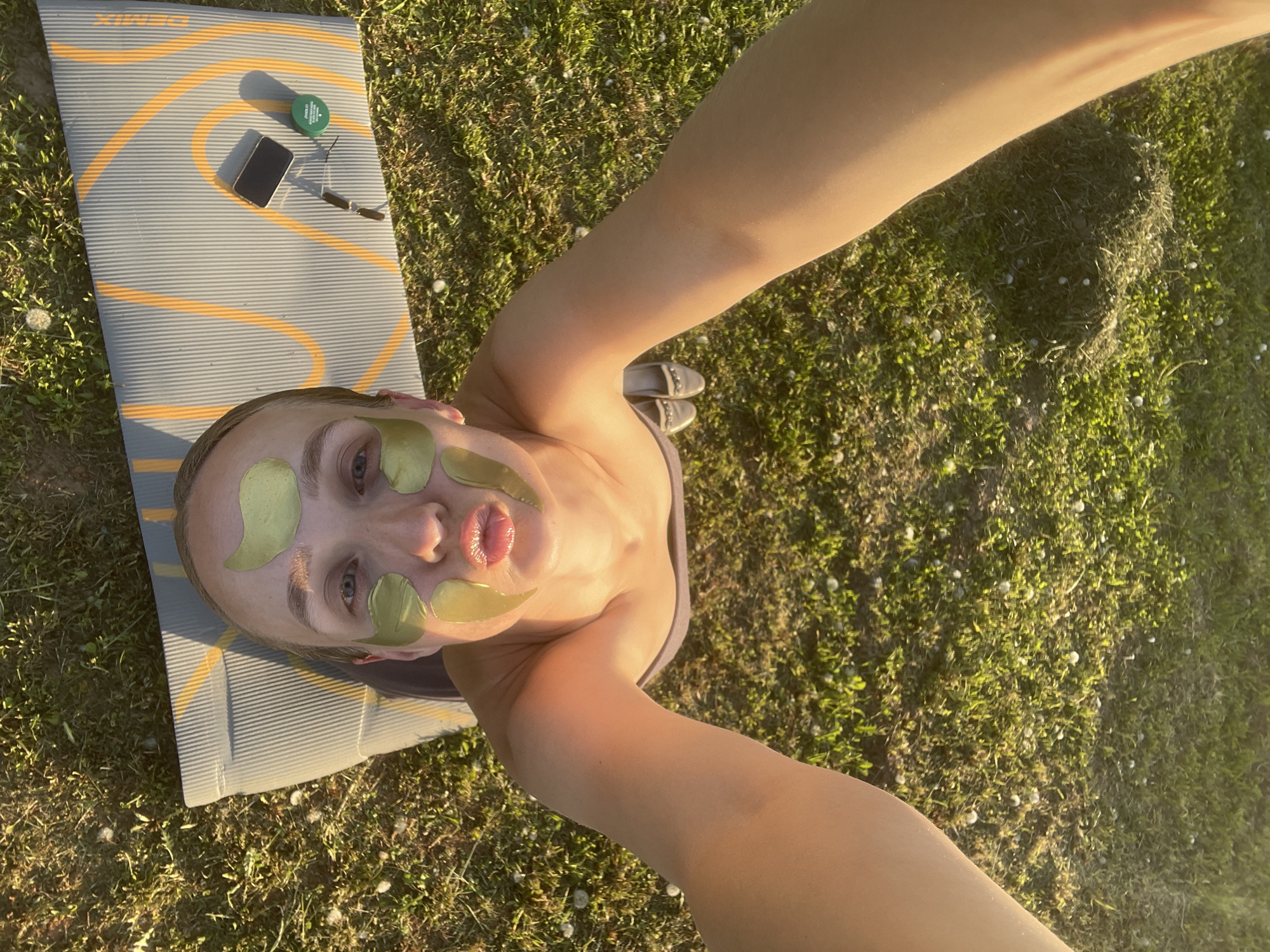 A woman wearing green eye and lip masks lies on a grey and orange yoga mat on a grassy area, possibly in Подшивалово, Russia. Her arms are extended towards the camera in a selfie pose. On the mat near her head are a green container, a cell phone, and a pair of sunglasses. She is also wearing flat shoes, and there are dandelions in the surrounding grass. The scene is lit by sunlight.