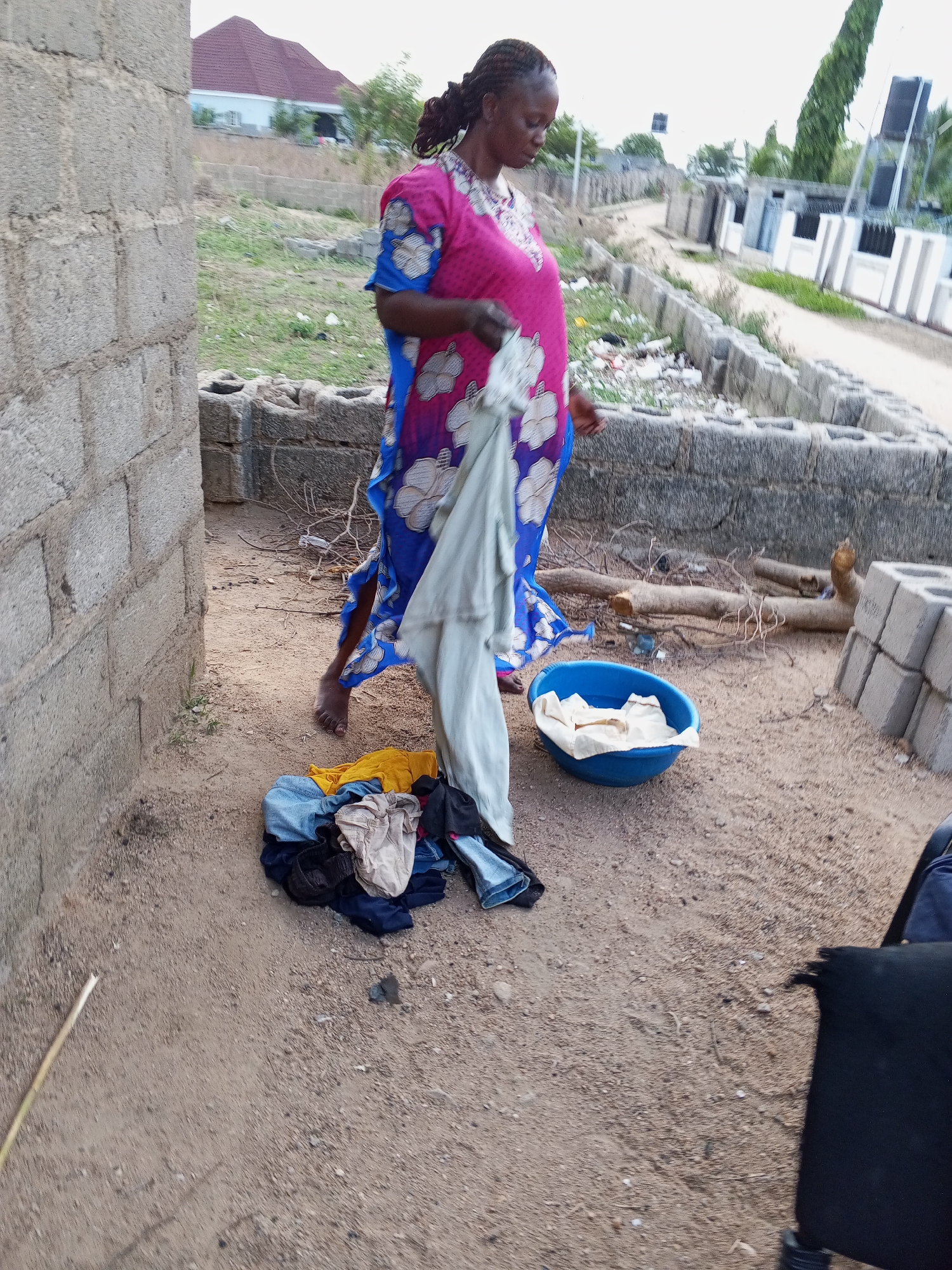 The image depicts a woman in a pink and blue floral dress standing in a dusty outdoor area in what appears to be Adonkolo, Nigeria. She is holding a light-colored piece of clothing. A pile of clothing in various colors lies on the ground near her bare feet. A blue plastic bowl contains more clothing. In the background, there is a low concrete block wall, a partially constructed brick building, and a street with houses. Other blocks of concrete are nearby. Some discarded debris is visible near the wall. The environment appears to be residential and under construction.