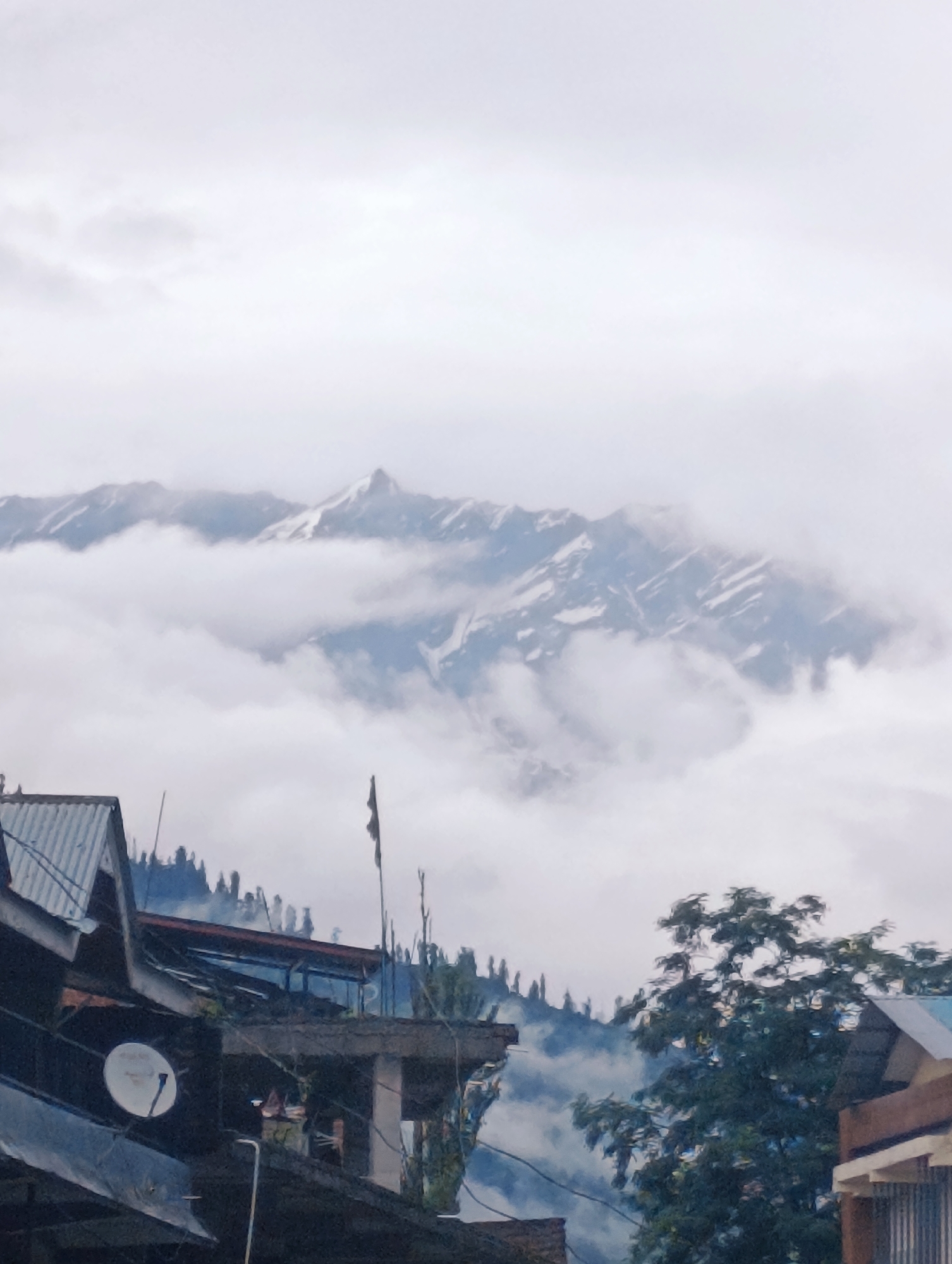 The image shows a panoramic view of a mountainous landscape, likely in Chichoga, India, on a cloudy day. In the foreground, there are several buildings with corrugated metal roofs and visible satellite dishes. A dense green tree stands to the right, partially obscuring the view of further structures. In the background, snow-capped mountains rise above a layer of thick white clouds. The sky is overcast, with diffuse light suggesting a humid or misty atmosphere.