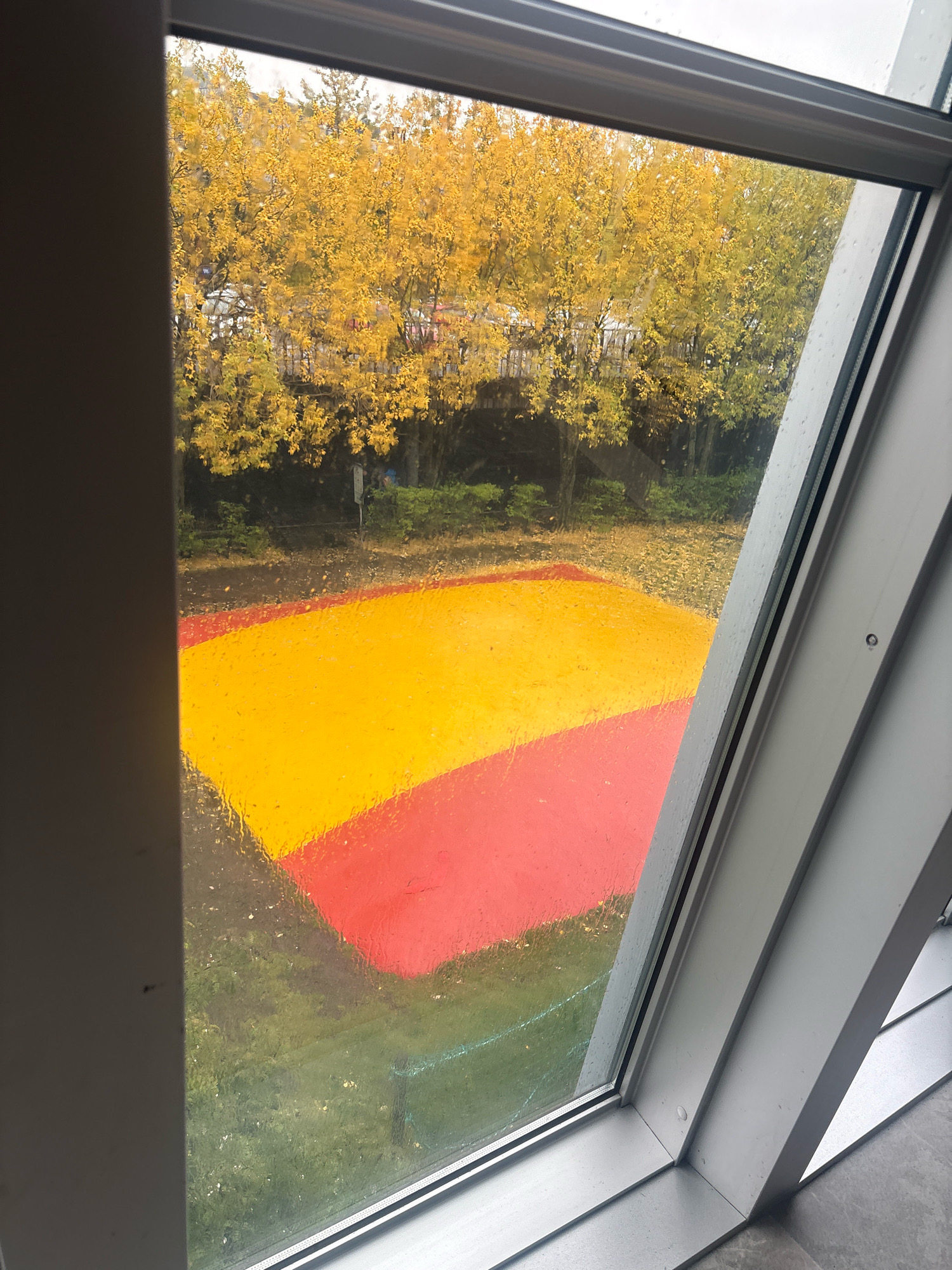 This is an exterior shot through a rain-streaked window, looking out onto an outdoor area. The scene depicts a rectangular patch of grass with a large, brightly colored geometric shape painted on it, divided into sections of red and yellow. In the background, trees with yellow autumn foliage are visible, suggesting a fall season. The window frame, made of silver-colored metal, is angled within the frame, indicating a slanted window or skylight. The setting appears to be an urban environment, possibly a park or public space in Reykjavik, Iceland, given the context. There are no people or animals visible in the frame.