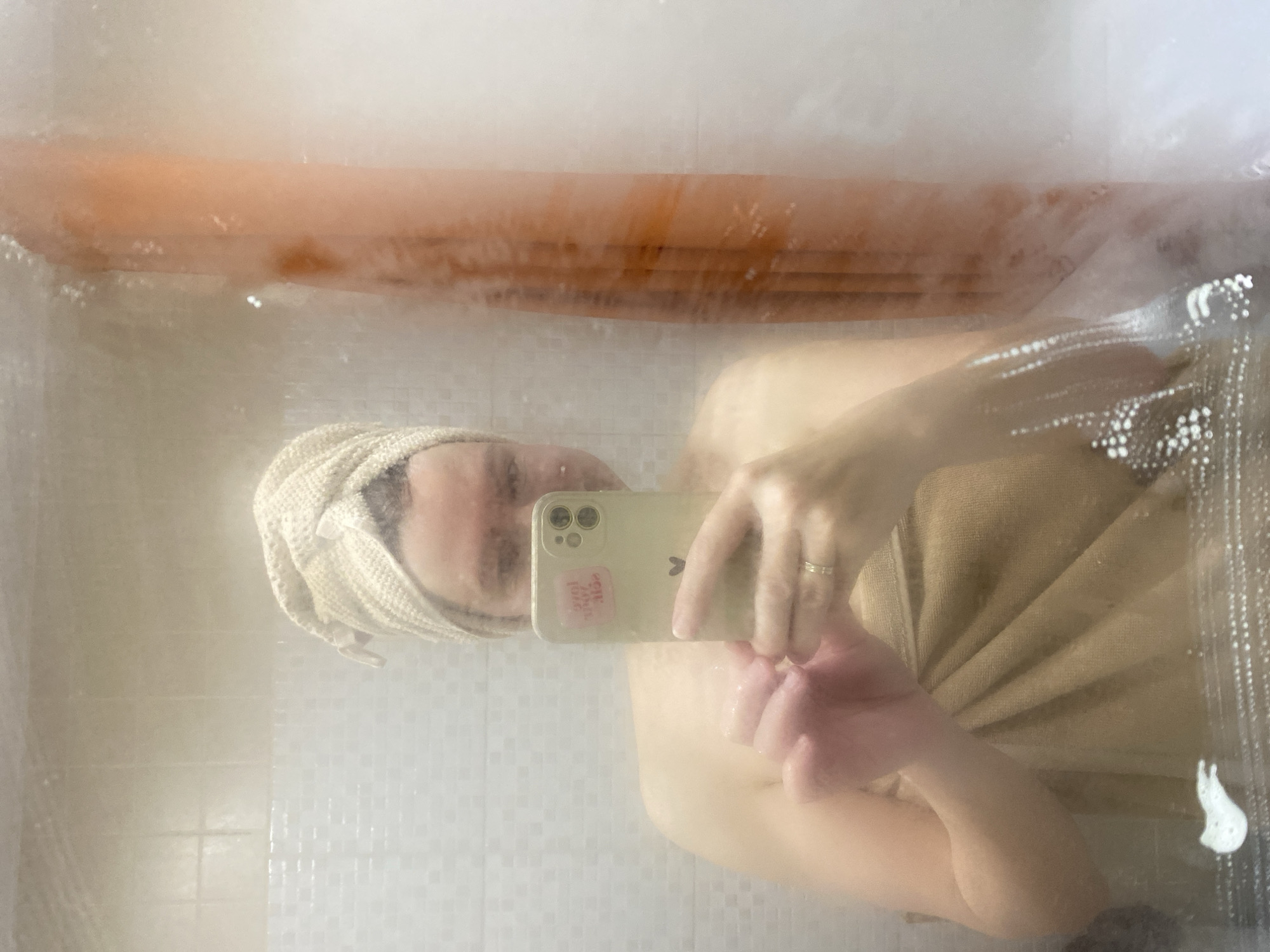 A person is taking a selfie in a steamy shower. The person has a towel wrapped around their head and is holding a smartphone in their hand. The phone has a sticker on the back. The person appears to be wearing a towel or wrap around their body. The shower has a glass door with condensation and streaks of water. A wooden slatted bath mat is visible at the top of the frame. The bathroom tiles are small and square. The location is Chaikovsky, Russia.