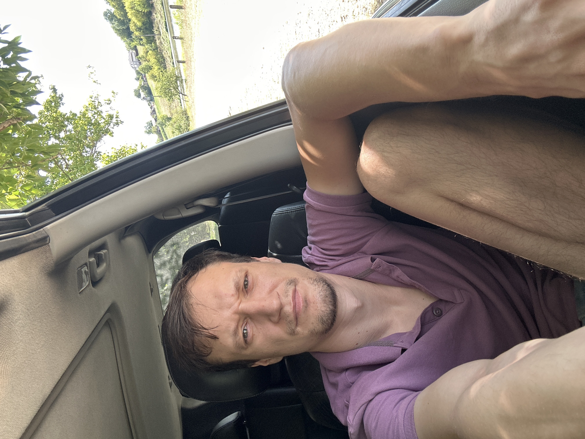 A man is sitting in the passenger seat of a car, looking at the camera with a slightly furrowed brow. He is wearing a short-sleeved purple polo shirt. His right arm is extended, resting on the open car door, and his legs are visible. The car appears to be parked outdoors, with a tree branch and foliage visible through the open window. The background shows a sunlit rural landscape with rolling hills, houses, and a body of water. The image is a selfie, taken from inside the vehicle, and it is daytime. The location is identified as Makiv, Ukraine.
