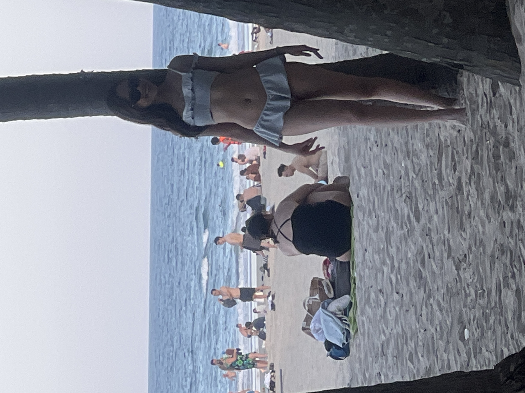 The image captures a scene on a sandy beach in Da Nang, Vietnam, with the ocean visible in the background. In the foreground, a young woman wearing a light blue bikini and sunglasses stands between two palm trees, facing the camera. Behind her, other beachgoers are scattered across the sand. Some are sitting or lying down, while others appear to be standing and interacting. The water is calm with small waves. Beach towels and bags are visible on the sand in front of some of the seated individuals. The overall atmosphere is that of a typical beach day.