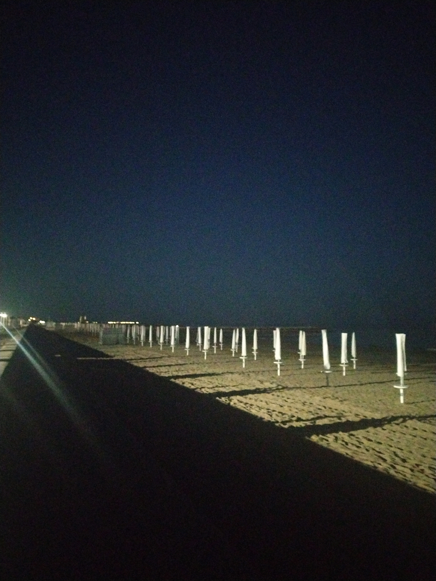 The image depicts a nighttime scene on a beach in Caorle, Italy. The sky is a deep, dark blue, with the horizon barely visible. The beach itself is sandy and appears to be sparsely populated. A row of white beach umbrella poles, closed and secured, stretches horizontally across the frame, casting long shadows on the sand. The poles are spaced evenly apart, suggesting an organized setup. To the left, a dark, paved walkway or pier runs along the edge of the beach, also casting a shadow. In the distance, faint lights suggest the presence of buildings or a town. The overall impression is one of quiet and stillness, with the beach deserted under the cover of night.