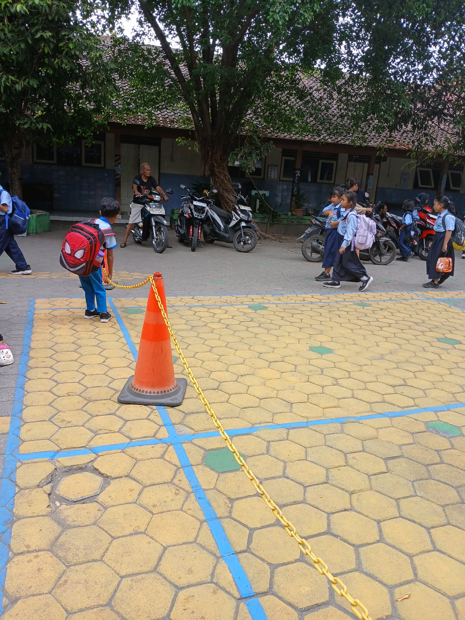 This image depicts a scene outside a school building, likely in Depok, Indonesia, given the context of the provided location. In the foreground, a young boy wearing a blue school uniform and a red Spider-Man backpack walks on a yellow hexagonal-patterned paved area. He is facing away from the camera, towards the right side of the frame. A bright orange traffic cone with a yellow chain around it is situated in the mid-ground, separating the paved area from the background.
In the background, several other students, mostly girls in blue and white school uniforms, are visible. Some are walking, while others appear to be near motorcycles parked in front of a low-slung building with a tiled roof. The building has several windows and appears to be a school structure. An adult man is also seen sitting on one of the motorcycles. Trees with dense foliage partially shade the area. The overall environment suggests a schoolyard or an area adjacent to a school, likely during a time of arrival or departure. The ground is paved with interlocking hexagonal stones of a yellowish hue, marked with blue lines indicating parking or pathways.