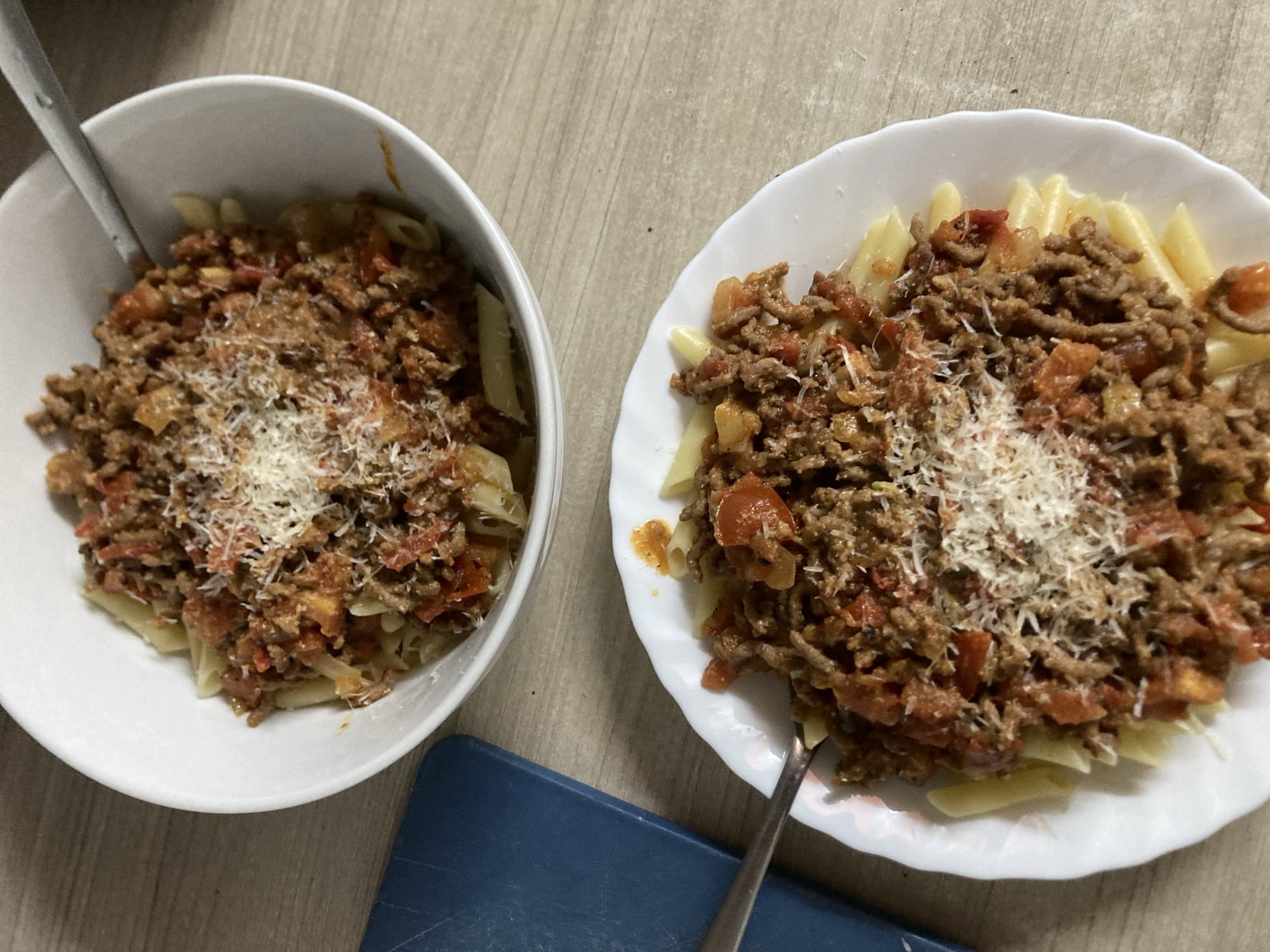 Two plates of penne pasta with bolognese sauce, topped with grated cheese, are arranged on a wooden surface. A blue cutting board is visible to the left of the upper plate. The lighting suggests an indoor setting, possibly a kitchen or dining area. There are no people or visible signs of activity in the image. The scene does not contain any elements that would indicate a specific location within the city of Barcelona, Spain.