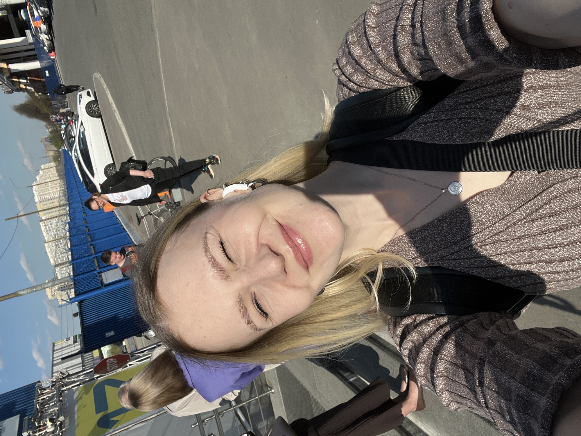 A woman with blonde hair and closed eyes is taking a selfie outdoors, wearing a brown ribbed top and a black backpack. She is wearing a silver necklace with a round pendant. In the background, there are blue shipping containers, a stop sign, and a white car parked on the side of the road. People are walking around, and there are buildings in the distance under a blue sky. The photo appears to be taken in a city environment, specifically in Khimki, Russia.