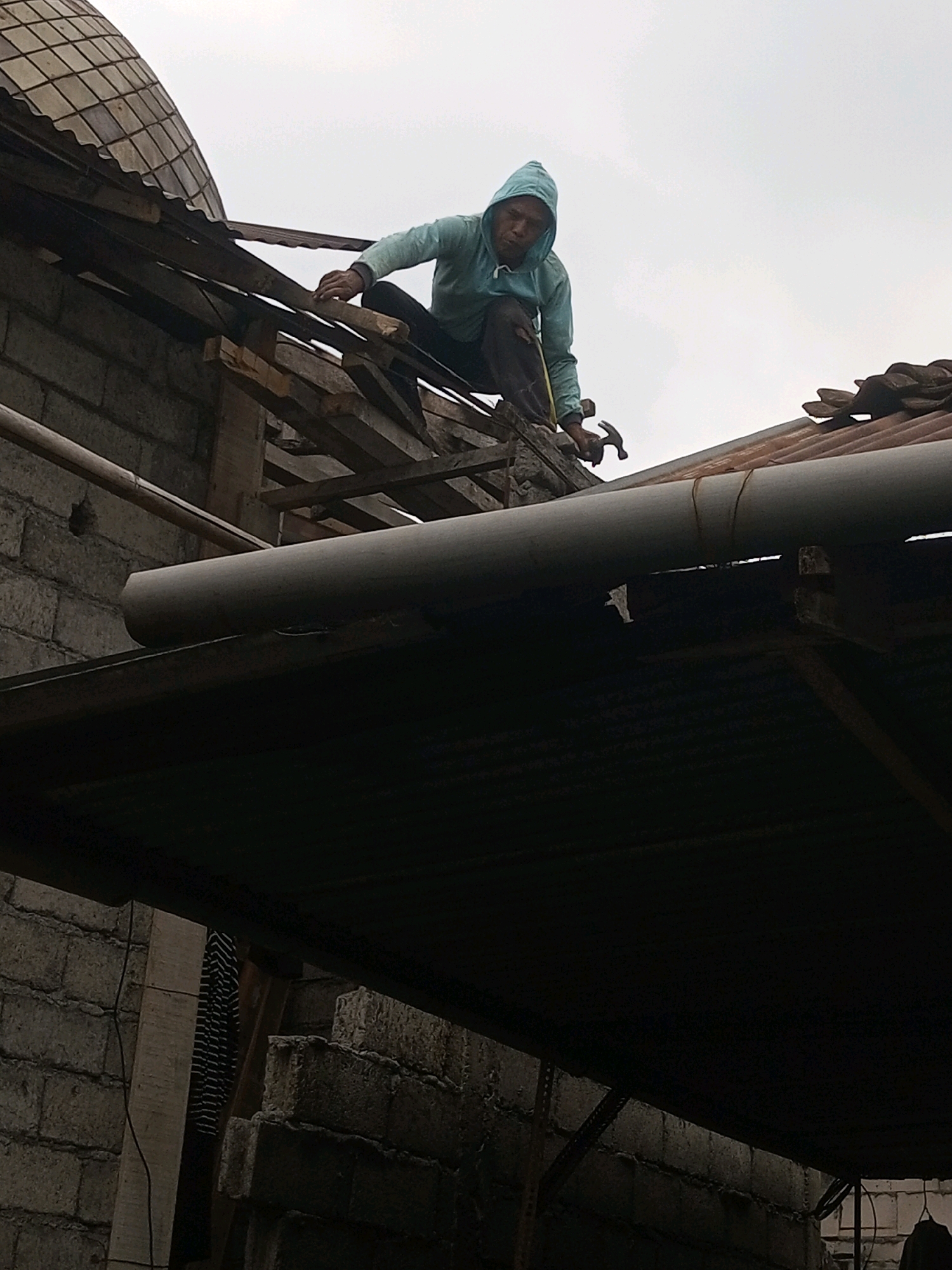 An Indonesian man in Andongsili, Indonesia, is working on a damaged roof. He is wearing a blue hooded jacket and dark pants, and is holding a hammer and a tape measure. The roof appears to be made of corrugated metal and tiles, and is supported by wooden beams. There is a large pipe running across the roof. The man is positioned on the roof, appearing to be measuring or securing a section of the roof. The surrounding environment consists of a brick wall structure and a cloudy sky.