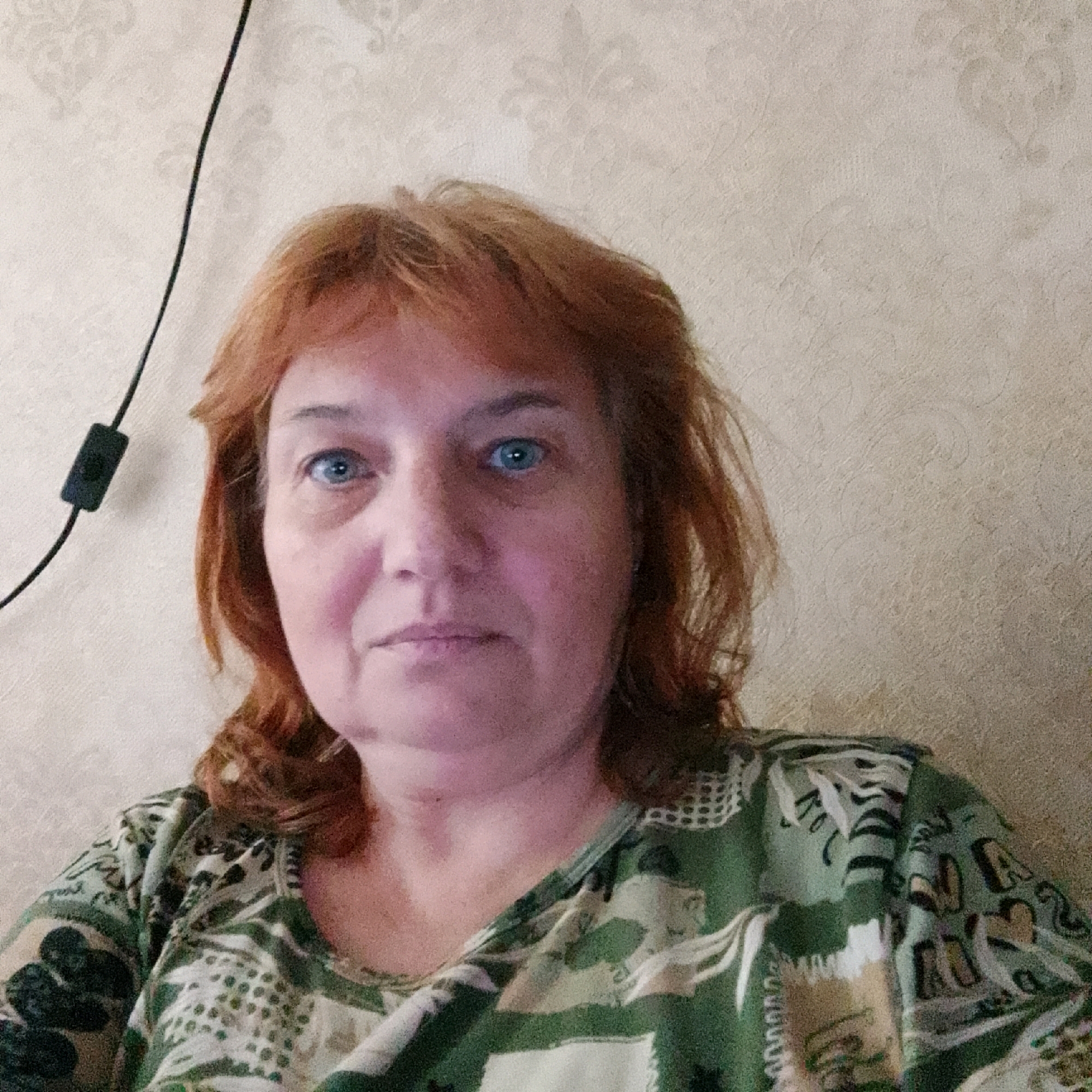 The image is a close-up, waist-up selfie of a middle-aged woman with reddish-brown hair, blue eyes, and fair skin. She is wearing a green and white patterned shirt. The background is a textured beige wall with a subtle floral pattern. A black electrical cord with an in-line switch is visible on the left side of the frame, running vertically. The lighting appears to be from an overhead source, casting soft shadows. The woman is looking directly at the camera with a neutral expression. The setting is indoors, likely a residential room. There is no specific indication of the location within the city of Narva, Estonia, as the image lacks any recognizable landmarks or environmental cues specific to that city.