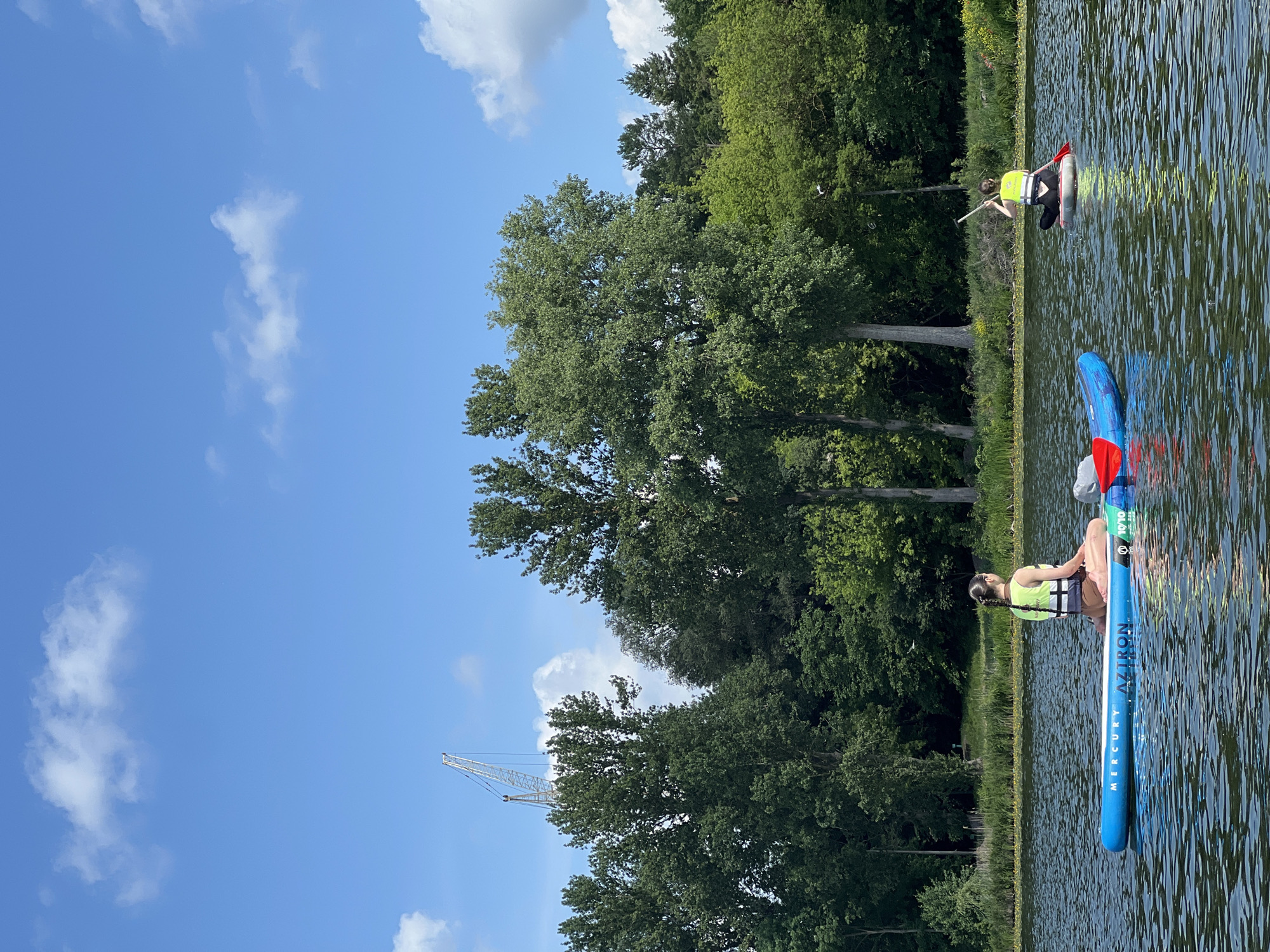 The image depicts a serene lake scene with two individuals on paddleboards. The person in the foreground is a woman with dark hair in a braid, wearing a neon yellow life vest and brown shorts. She is seated cross-legged on a blue "MERCURY AZTRON" paddleboard, facing away from the camera. A red and blue paddle rests on the board beside her. In the background, another person, also wearing a neon yellow life vest, is seated on a white paddleboard and paddling with an oar.
The lake is calm, with gentle ripples on the surface. The shoreline is lined with lush green trees, forming a dense forest. The sky is a vibrant blue with scattered white clouds. In the distance, a large construction crane is visible above the treeline, suggesting the presence of development or industrial activity within or near the city of Bila, Ukraine. The overall atmosphere is peaceful and recreational, with the natural beauty of the lake and surrounding forest contrasting with the man-made structure in the background.