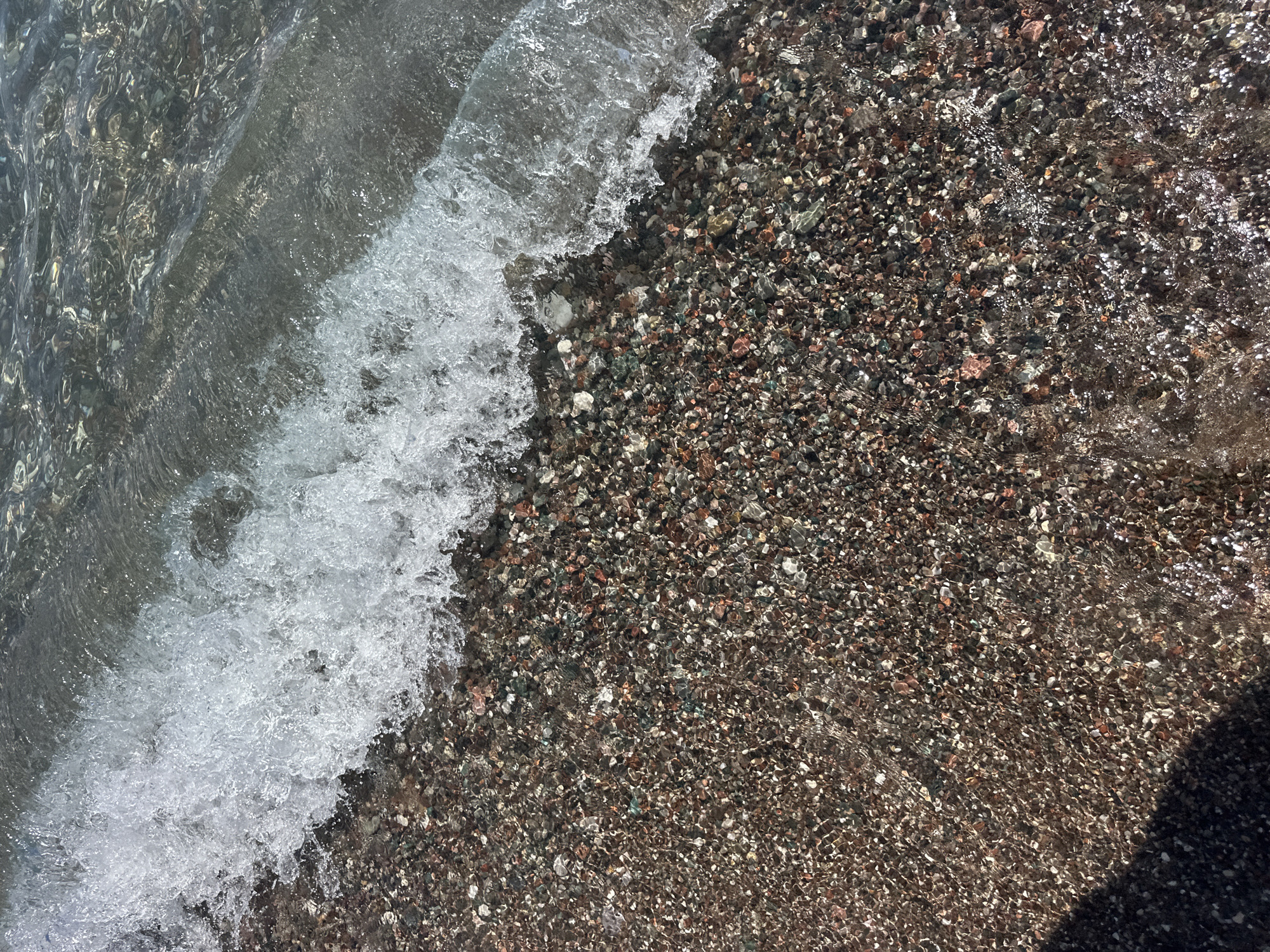 The image captures a close-up view of a pebble beach with a gentle wave breaking on the shore. The pebbles are small and varied in color, ranging from dark brown and gray to hints of red and white, many of them appearing wet and glistening. The water is clear and reflects the sunlight, creating shimmering patterns on the wet pebbles. The wave is white and foamy, appearing to be in the process of receding. The overall scene depicts a natural coastal environment. The location is identified as Boreti, Montenegro.