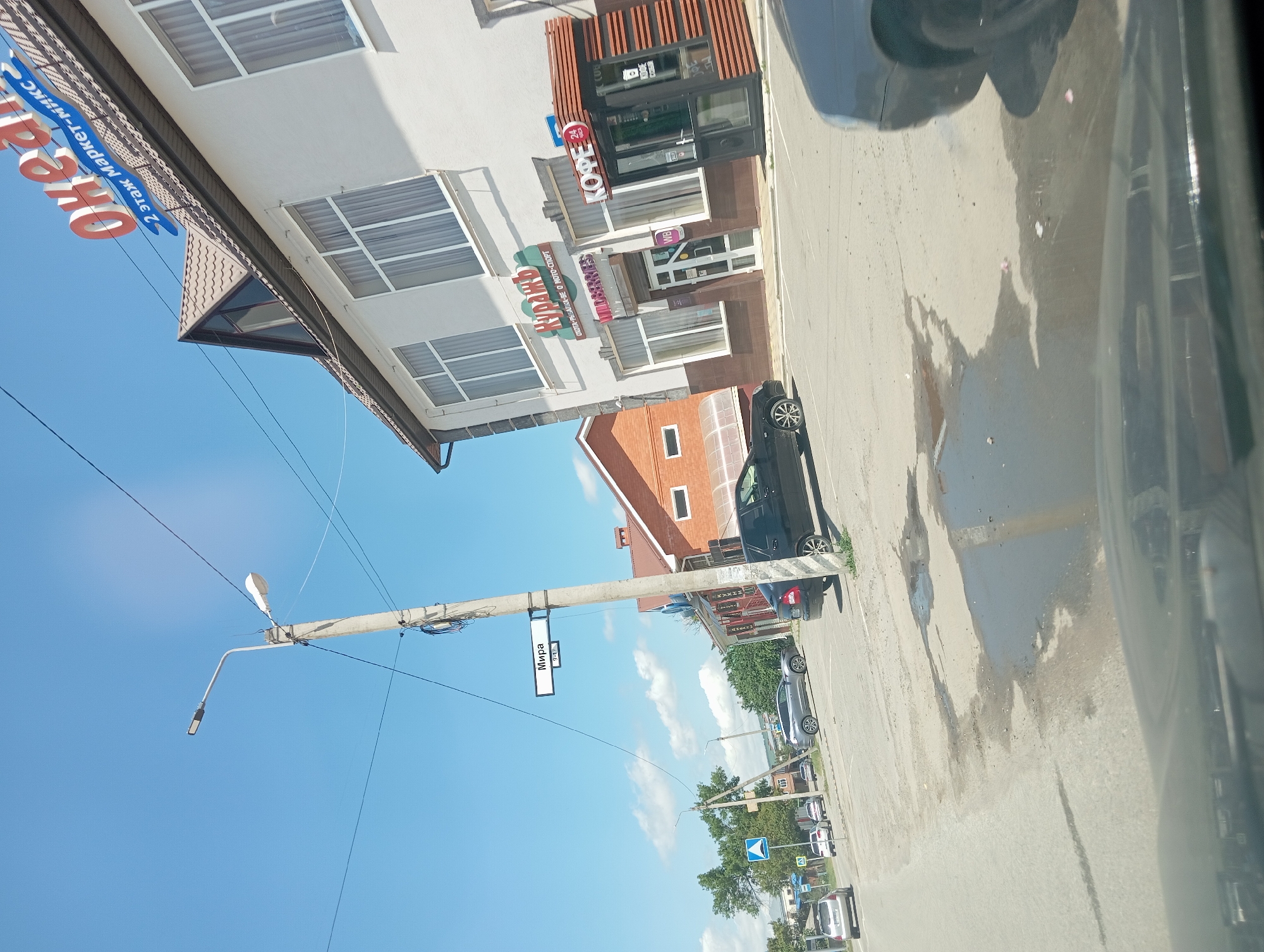 The image depicts a street scene in the city of Otradnaya, Russia, under a clear blue sky with scattered clouds. A concrete lamppost stands prominently in the foreground, with electrical wires running across it and into the sky. A street sign attached to the lamppost reads "Мира" (Mira) with numbers "8-17" below it, indicating a street name and house number range.
To the right of the lamppost is a white building with a dark brown lower facade and a brown-tiled roof. A large red and blue sign on the roof reads "Океан" (Ocean) with smaller text below it in blue that says "2 этаж Маркет-микс" (2nd floor Market-mix). On the white facade of this building, there are several large windows with white frames and light-colored curtains. Below some of the windows are signs for businesses: one in green text says "КУРАЖ" (Kurazh) with additional text below in smaller font "ОХОТА-РЫБАЛКА-ОДЕЖДА-СПОРТ" (Hunting-Fishing-Clothing-Sport). Below that, a sign reads "wildberries". To the right of the "wildberries" entrance, there is a small kiosk-like structure with a sign above it that says "КОФЕ 24" (Coffee 24) and displays a red "24" icon.
In front of the building, a black car is parked. Further down the street, a black sedan is visible. Other cars are present in the background, including a silver SUV and a silver hatchback. A traffic sign with a blue background and white symbols is visible on the left side of the road. The road surface is asphalt with several large puddles and some scattered debris. Trees and other buildings are visible in the distance, suggesting an urban or semi-urban environment. The image appears to have been taken from inside a vehicle, as the bottom of the frame shows the reflection on a car's dashboard or windshield.