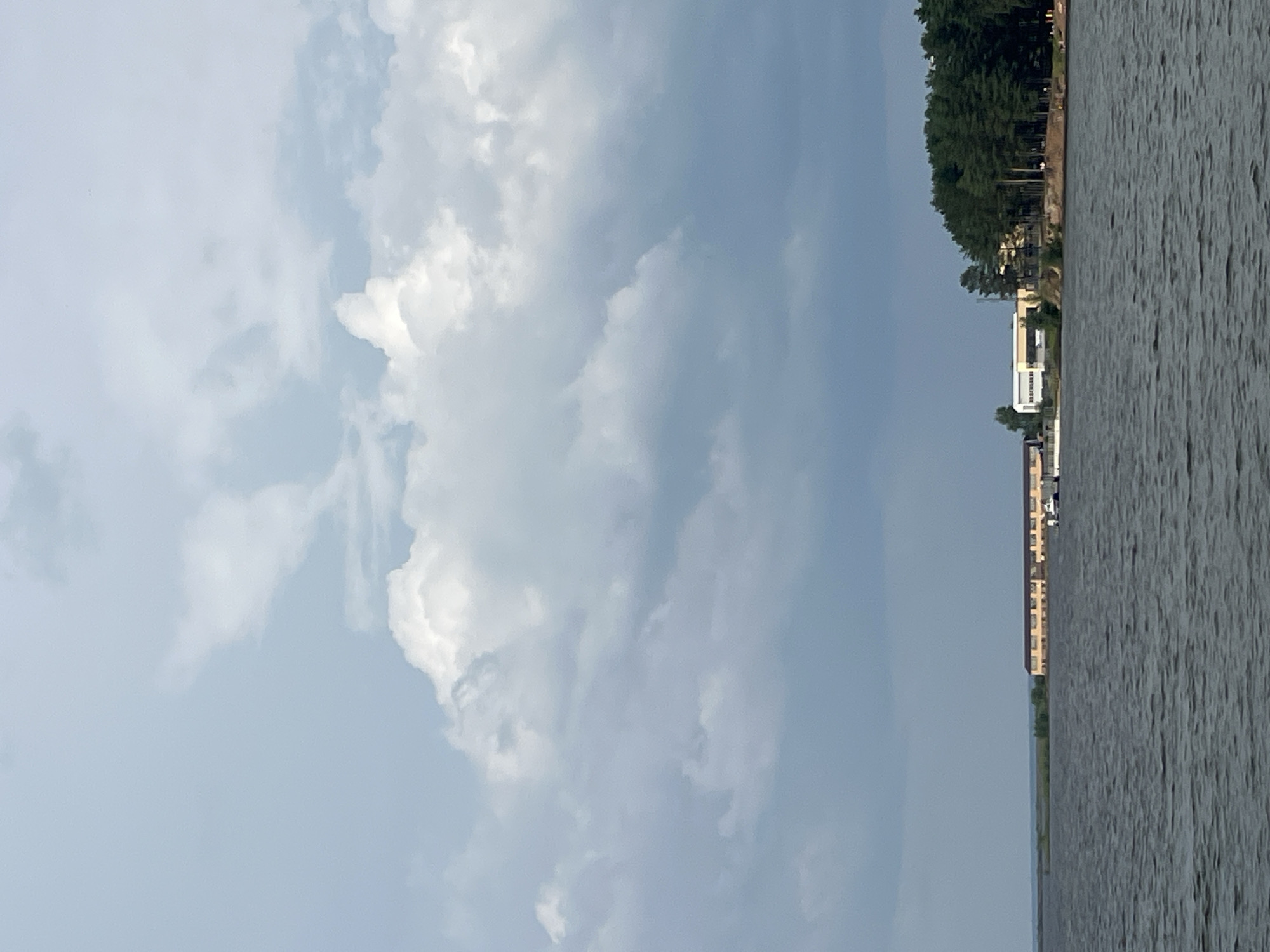The image depicts a body of water, likely a reservoir or large river, with buildings and a dense forest lining the far shore. The sky is filled with dramatic, cumulonimbus clouds, indicating potential for inclement weather. In the foreground, the water is dark and rippled, suggesting a moderate wind. Along the shoreline, several structures are visible. To the left, a long, light-colored building with multiple windows runs parallel to the water. To its right, a cluster of buildings appears to be part of a resort or industrial complex, with one building featuring a distinctive white roof. A line of tall pine trees forms a dense backdrop behind these structures, creating a stark contrast with the open sky and water. The scene is likely captured from a boat or the opposite shore, offering a panoramic view of the water and the developed coastline. The location is identified as Chaikovsky, Russia, a city known for its hydroelectric power station and reservoir on the Kama River.