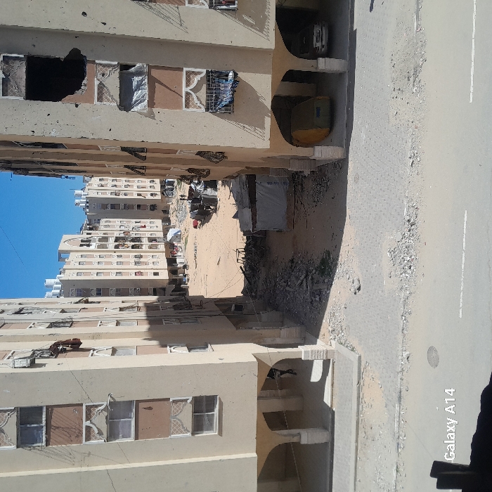 The image depicts a damaged urban residential environment in Khan Yunis, Palestinian Territory, under a clear blue sky. Two multi-story, light-colored apartment buildings frame a central, unpaved area. The building on the right exhibits significant structural damage, including large impact craters on upper floors and shattered windows, some covered with fabric or plastic. The building on the left shows some obscured windows. Both buildings feature ground-level arched passages. Under the right building's arches, a yellow object (possibly a vehicle) and a silver car are parked. One person, dressed in dark clothing, is visible walking under the left building's arches.
The central area between the buildings is an expansive dirt and sand lot strewn with debris, rubble, and what appear to be makeshift shelters constructed from tarpaulins and various materials. A bicycle lies on its side amidst the debris. Further into the dirt area, a white tent is visible, and several small figures, likely people, are present near it in the distance. Additional multi-story residential buildings, some with rooftop water tanks, are visible in the background, surrounding the central lot. The foreground shows a partially paved, debris-strewn ground leading into the unpaved area. The image captured by a "Galaxy A14" camera.