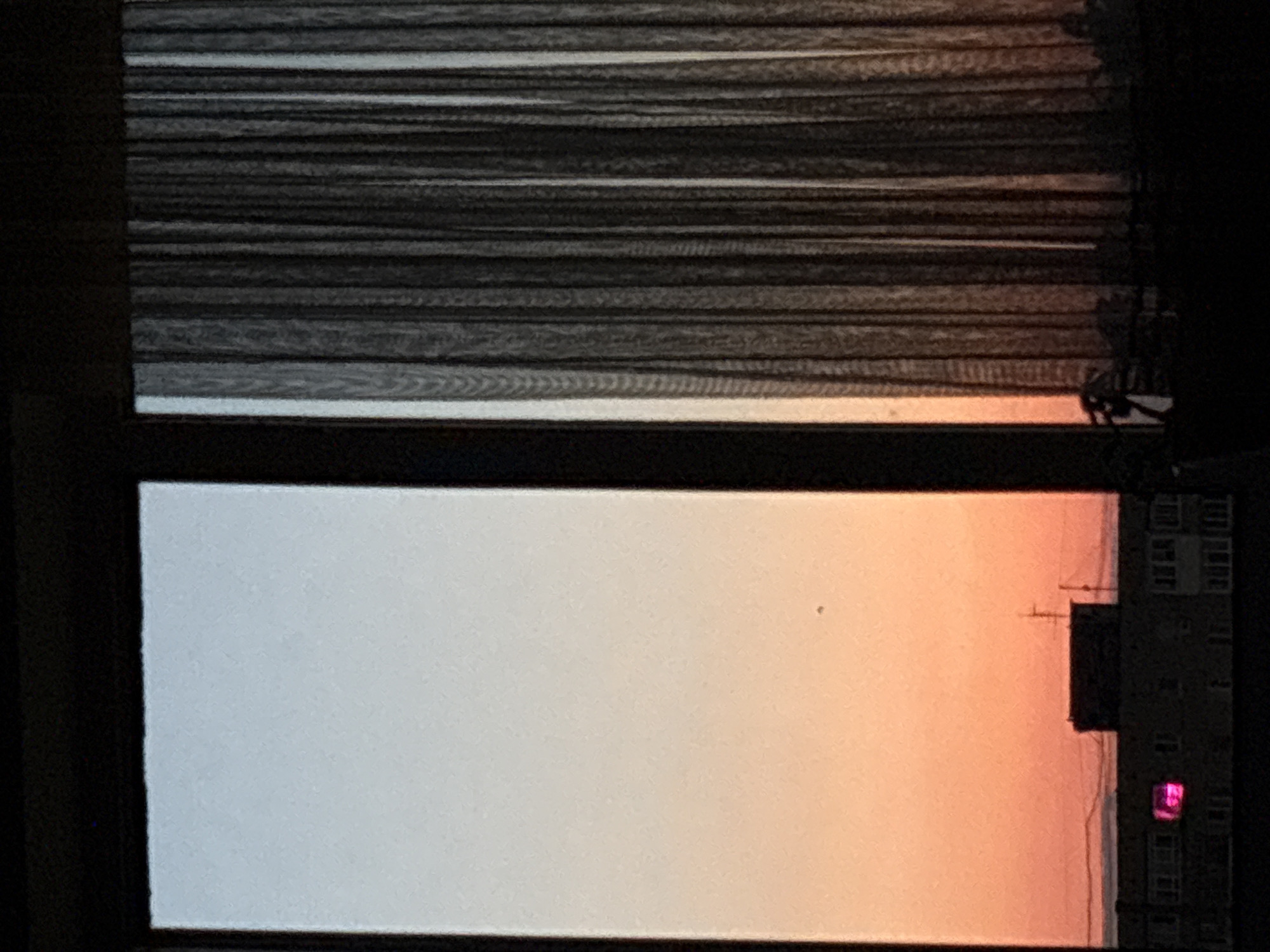 The image depicts a view through a window. Venetian blinds are visible at the top of the frame. Below the blinds, the window reveals a sunset sky transitioning from white to orange. A building with multiple windows is visible on the right side of the frame. One window is illuminated in pink. The exact location in Chaikovsky, Russia is not possible to determine from the given image.