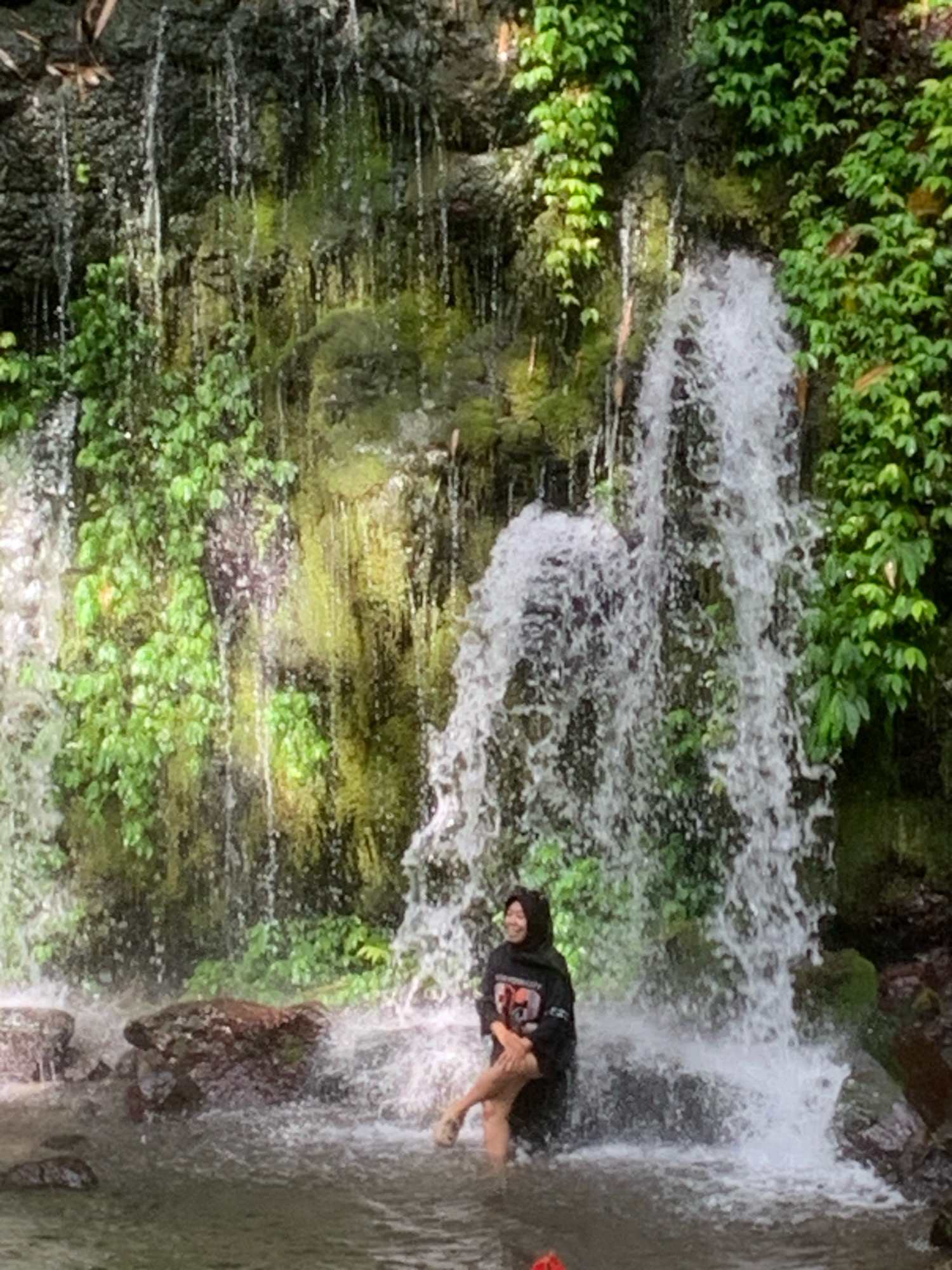 A young woman wearing a black hijab and a black hoodie with a graphic on the front is sitting with her legs crossed in the shallow water at the base of a waterfall. She is smiling and looking to her left. The waterfall cascades down a moss-covered rock face surrounded by lush green foliage. The water is clear and appears to be flowing over rocks and into a pool where the woman is seated. The scene is located at a waterfall in Pronojiwo, Indonesia, a region known for its natural beauty and numerous waterfalls.