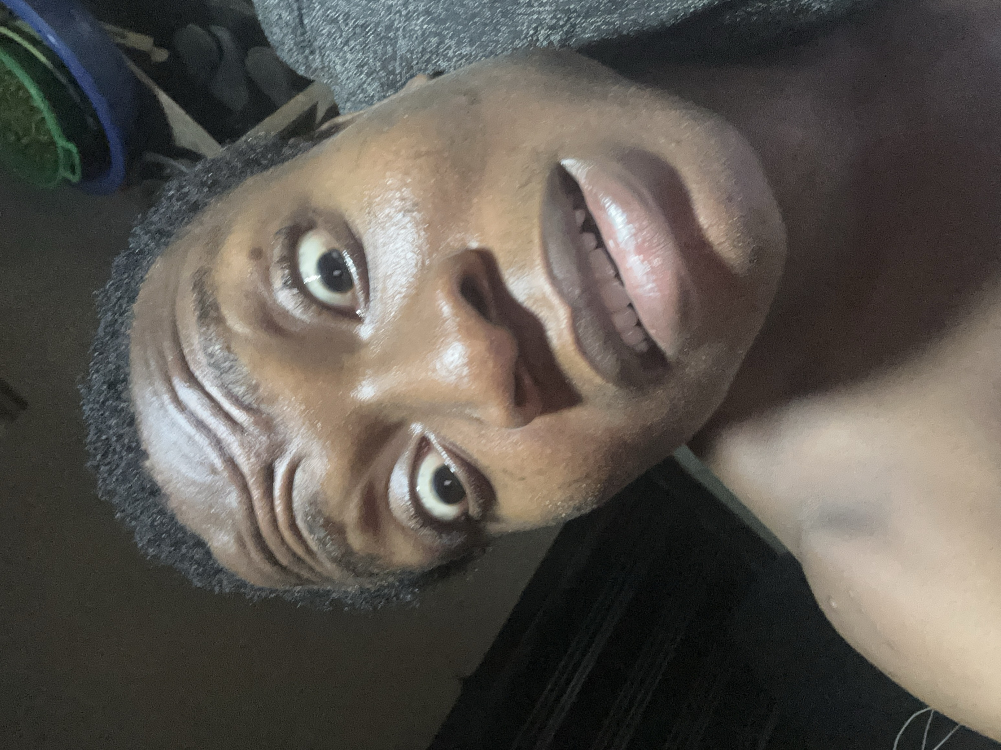 A close-up, angled view of a person's face. The person has dark skin, short curly hair, and is looking directly at the camera. The forehead has visible wrinkles, and the face appears to be slightly shiny. The lips are parted, revealing teeth. The background is dark and out of focus, but there's a hint of clothing or objects in the upper left corner. The location and specific details about the environment are unknown.