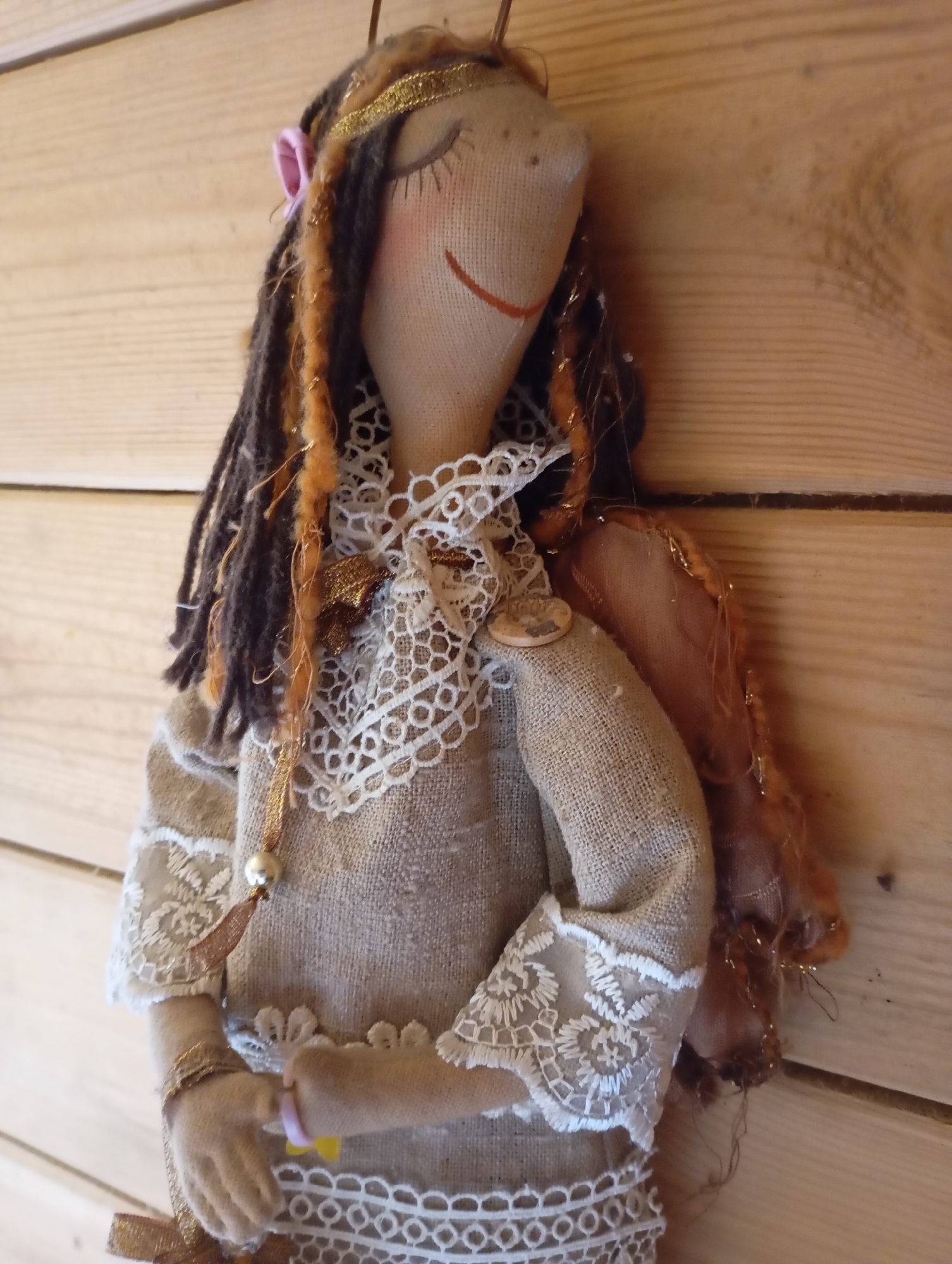 This is a close-up, eye-level, medium shot of a handmade fabric doll. The doll is standing against a background of horizontal wooden planks. The doll has a neutral-colored, burlap-like fabric body and is dressed in a similarly textured garment adorned with white lace trim. It has braided hair, some of which is dark brown and some is a coppery orange, interwoven with gold thread. The doll has closed eyes, depicted with eyelashes, and a red stitched smile. A pink ribbon is visible on its head, and a gold ribbonband goes across its forehead. It wears a lacy white collar and has thin fabric wings on its back. A gold chain with a large pearl hangs from the neck area, and the doll's hands are clasped together, holding a small pink ring and a yellow object. A gold bow is tied around its waist. The overall aesthetic of the doll suggests a whimsical, perhaps bohemian or fairy-like character. The lighting is soft, suggesting it might be indoors with ambient light, or outdoors on an overcast day. There is no visible text in the image. The setting appears to be a rustic interior, given the wooden plank background.