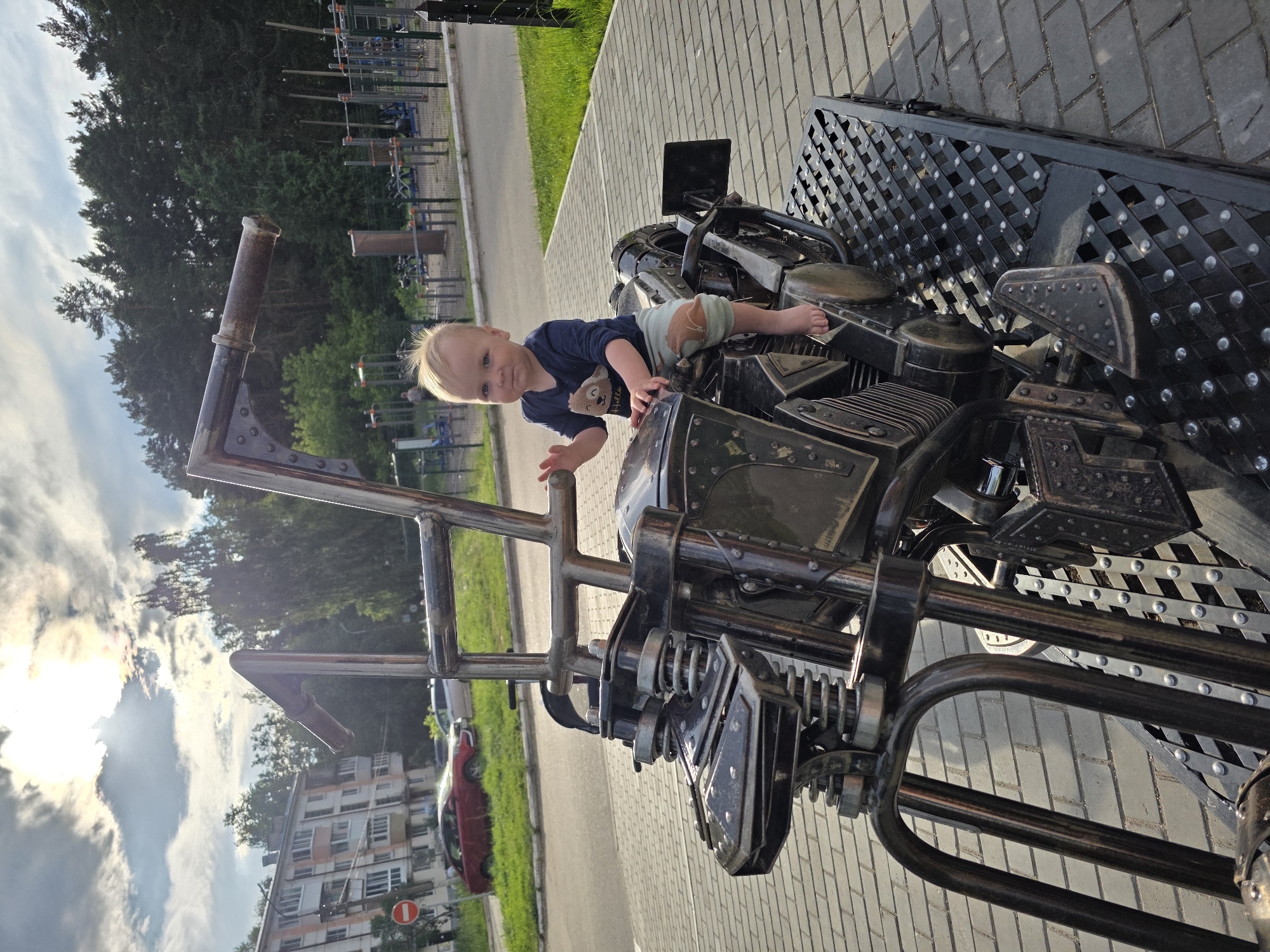 A Caucasian toddler with blonde hair, wearing a dark blue shirt with a bear graphic and light green shorts with a patch, sits on a metal sculpture of a motorcycle. The toddler is barefoot and looking at the camera, with their right hand raised as if waving. The motorcycle sculpture is black with silver accents and is positioned on a metal platform. The background shows a street with a red car, a multi-story apartment building, trees, and a park with outdoor exercise equipment. The scene is outdoors on a paved area with a brick pattern. The lighting suggests it is daytime, possibly late afternoon, with the sun partially obscured by clouds. The location is identified as Chaikovsky, Russia.
