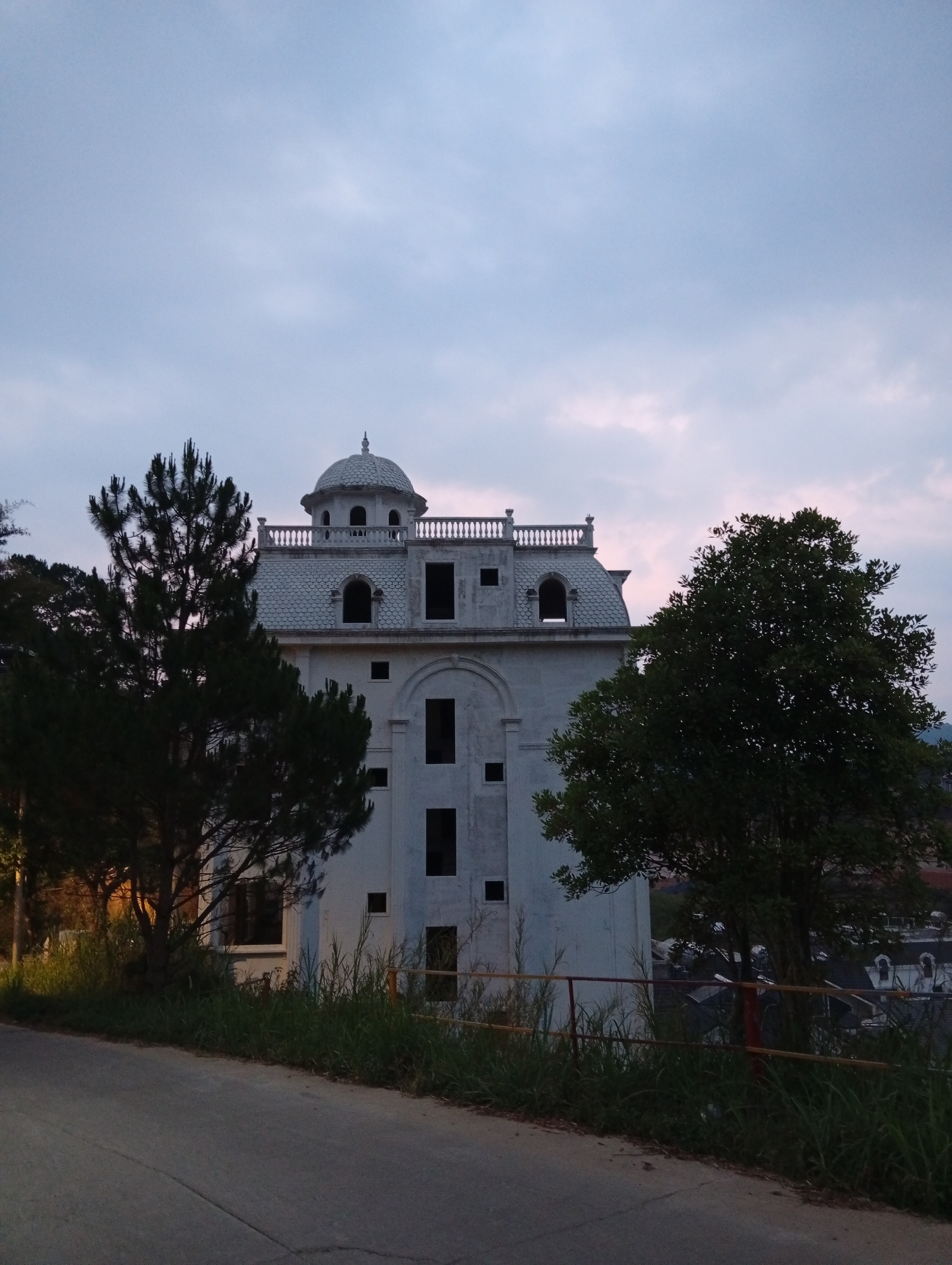 The image shows a tall, unfinished white building with architectural details like a dome, arched windows, and a balustrade. It appears to be in Da Lat, Vietnam, based on architectural style and vegetation. The building lacks window glass. It stands on a slope above a road with a yellow and red guardrail. Two trees, one evergreen and one deciduous, frame the building. In the background, glimpses of densely packed buildings are visible. The sky is overcast with hues of blue and pink.