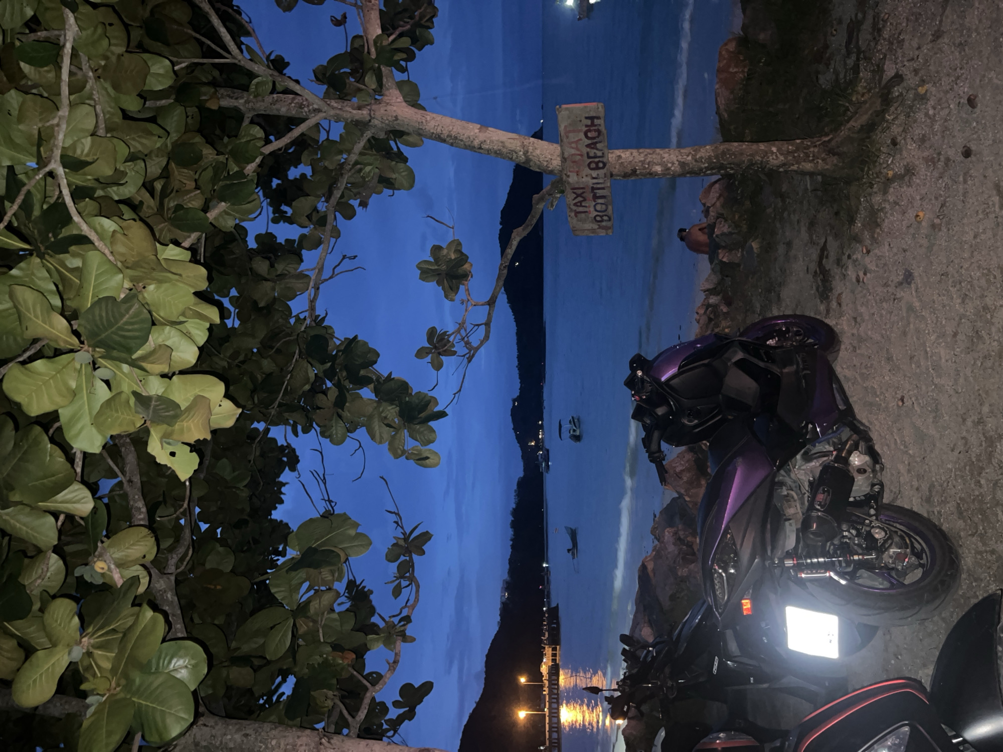 The image depicts a twilight scene on a beach in Ko Pha-ngan District, Thailand. In the foreground, two scooters are parked on a sandy and rocky area. The scooter on the right is purple and black, with visible engine parts and suspension. The scooter on the left is partially visible and appears to be black and red.
Behind the scooters, the sea stretches out, with gentle waves reaching the shore. Several boats are scattered across the water, and a pier with lights is visible in the distance to the left. The sky is a deep blue, transitioning from dusk.
To the right, a tree stands near the edge of the beach, with a weathered sign attached to its trunk that reads "TAXI BOAT BOTTLE BEACH". A single person is seated on rocks near the water's edge, facing the sea. Trees with large green leaves frame the top of the image, casting shadows and obscuring parts of the sky. The overall atmosphere suggests a peaceful evening on a tropical island.