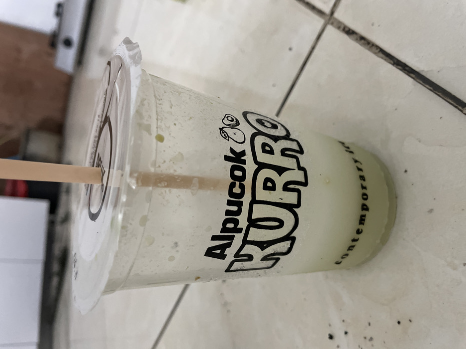 A partially consumed clear plastic beverage cup, branded "Alpucok KURRO" with an avocado icon and "contemporary ice" printed in black text, rests on a light-colored tiled floor with dark grout lines. The cup contains a low level of light green and white liquid, consistent with an avocado-based beverage, exhibiting small green particulate matter and exterior condensation. A beige drinking straw is inserted through the clear plastic dome lid. Dark specks of debris are scattered on the surrounding tiles. The background is out of focus, depicting indistinct indoor objects, consistent with a domestic or commercial interior in Kayen, Indonesia. No individuals are present.