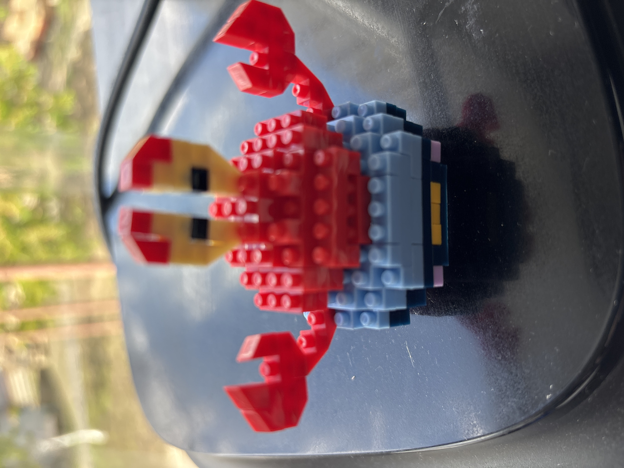 A small, toy crab, constructed from interlocking building blocks (resembling nanoblocks), is the central subject of the image. The crab is primarily red, featuring large red claws and multiple red legs. Its body section is light blue, and its head/eye area is yellow-gold with small black accents.
The toy is positioned on a dark, glossy, and slightly curved surface. This surface is reflective, showing a blurred outdoor background. The reflections prominently display bright green foliage and a light blue sky, suggesting the photograph was taken during the daytime, likely under bright or sunny conditions. Some dust or small specks are visible on the reflective surface, and a faint, blurred red reflection of the toy itself can be seen. The exact setting is ambiguous, but the appearance of the surface and reflections could indicate a car dashboard, a reflective window sill, or a similar indoor-outdoor boundary. There is no discernible activity or event taking place, beyond the static placement of the toy for the photograph. No people are visible, and there is no legible text in the image.