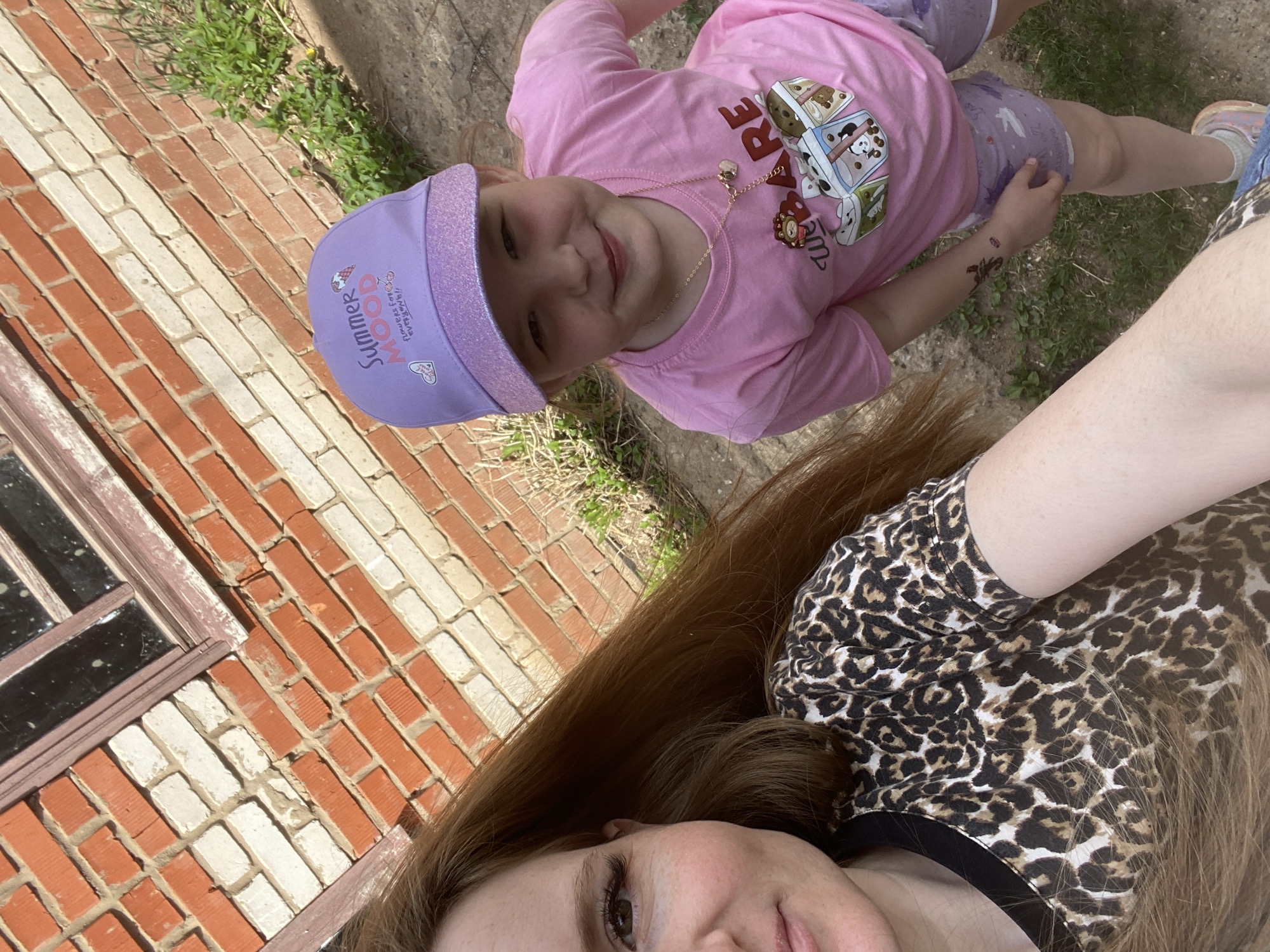 A photo, taken at a canted angle, depicts a fair-skinned woman and a young girl outside next to a brick wall. The woman has auburn hair and wears a leopard print top. The young girl is wearing a pink t-shirt with cartoon images, printed purple shorts, and a purple visor hat with "Summer Mood" printed on it. A gold necklace adorns her neck and a temporary tattoo is visible on her arm. They are on a patch of grass and dirt. Part of a red-brick building with window panes is visible in the background. The location could be near the city of Chaikovsky, Russia, based on the provided information.