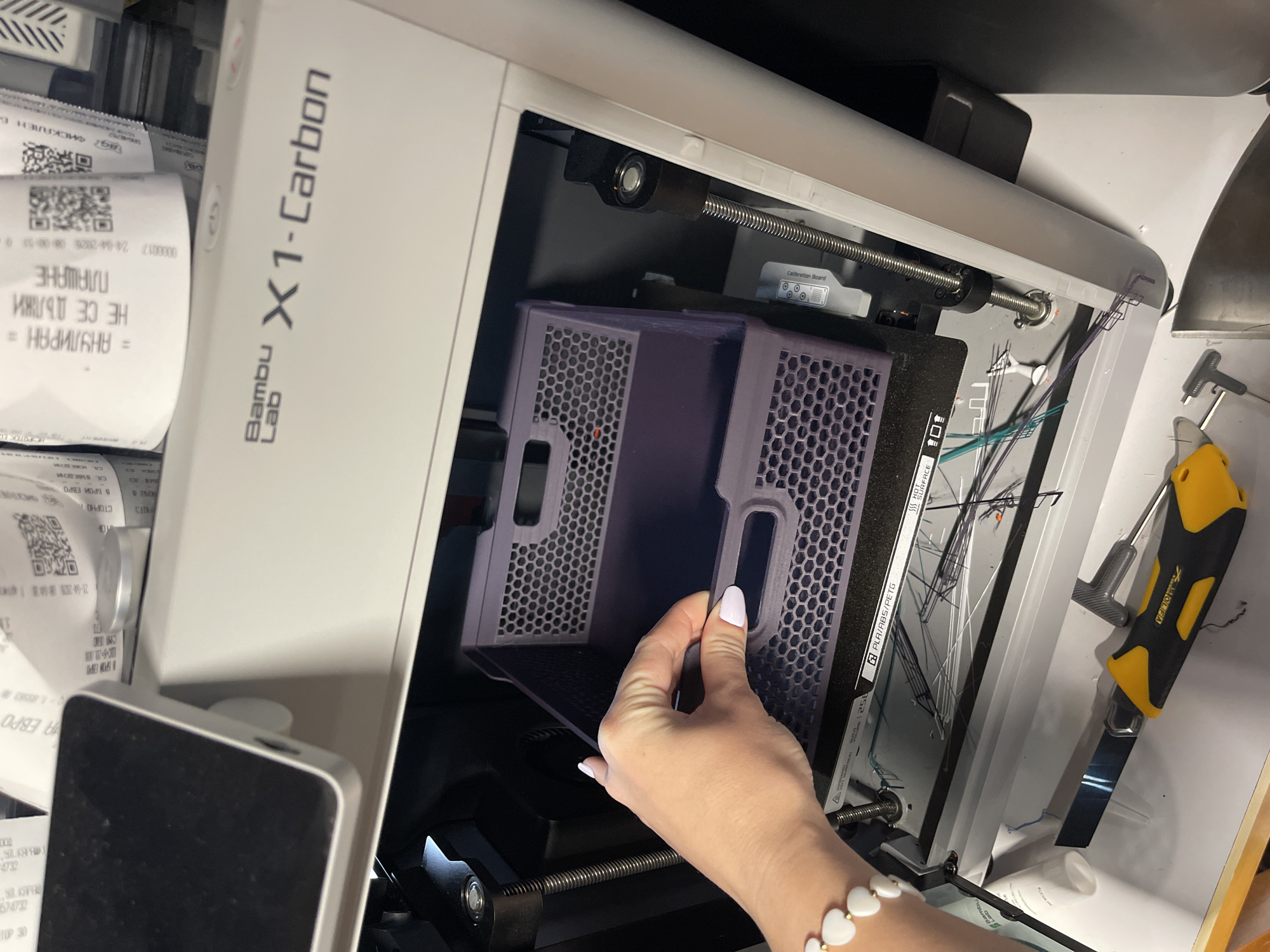 Here's a matter-of-fact description of the image:
**Scene:** Close-up view of the interior of a Bambu Lab X1-Carbon 3D printer.
**People:** A person with pale skin and manicured nails is inserting or removing a purple air filter element. They are wearing a white beaded bracelet.
**Subjects:** The main subjects are the 3D printer itself and its internal components, including the air filter.
**Objects:** Visible objects include the air filter, calibration board, metal guide rods, tools (utility knife and T-handled Allen wrench), waste filament strands, a bottle of grease, and receipts.
**Actions:** The action shown is the person handling the air filter.
**Interactions:** There is interaction between the person and the 3D printer's air filter.
**Environment:** The environment appears to be a workshop or lab setting, given the tools and receipts strewn around.
**Specific details:** Some receipts display Bulgarian Cyrillic script. The receipt also shows "Sofia".