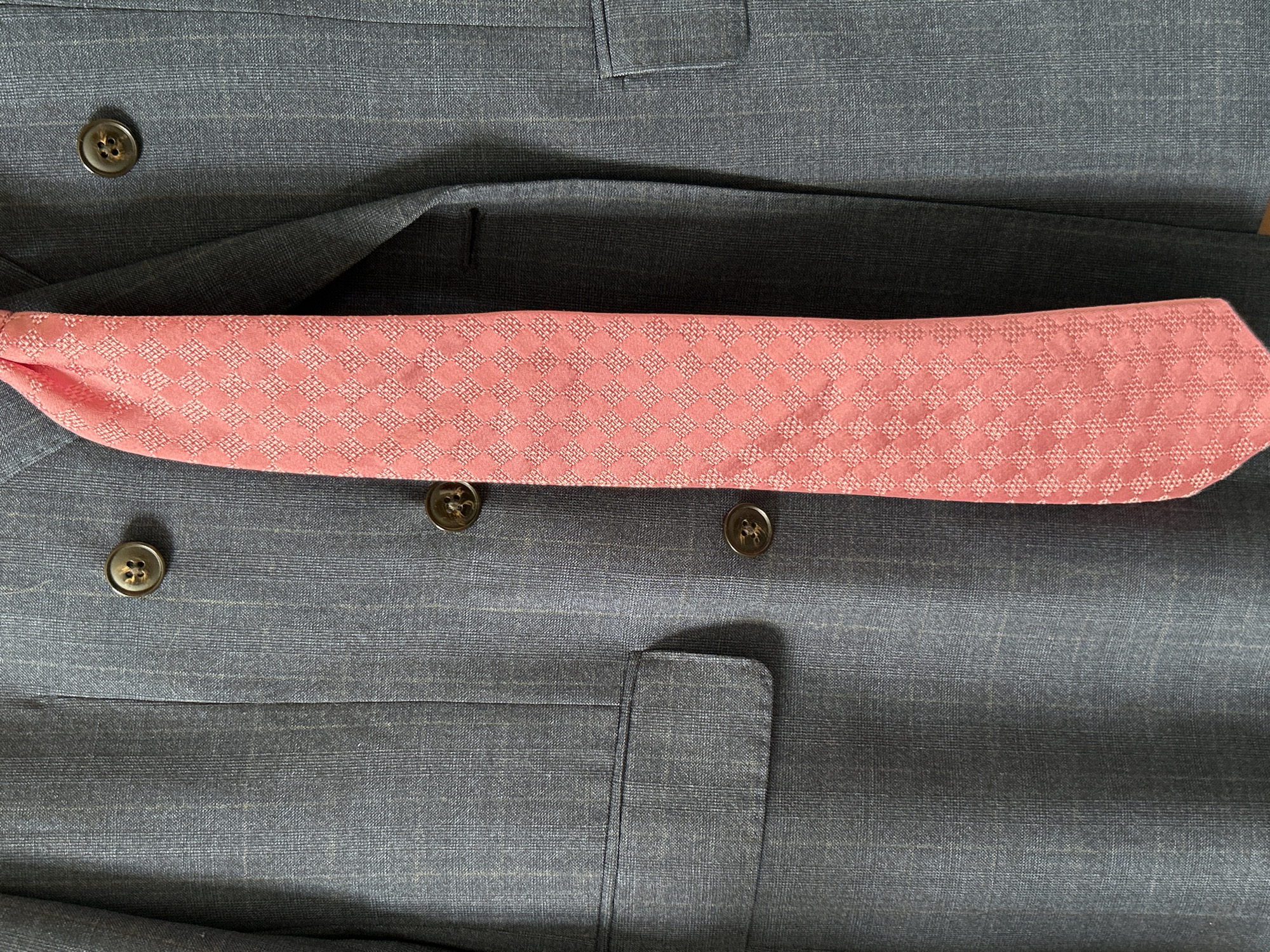 A close-up, vertical, eye-level shot shows a portion of a dark grey pinstripe suit jacket and a bright pink patterned tie. The jacket has four dark brown buttons visible, two in a double-breasted arrangement, and a flap pocket on the left side. The tie is positioned vertically on the right side of the jacket, partially obscuring the buttons. The fabric of the suit is textured with faint vertical pinstripes. The tie features a repeating diamond pattern with a lighter pink weave. The lighting appears to be natural, casting subtle shadows. The background is entirely filled by the suit jacket and tie. The scene is indoors, and no other objects or people are visible.