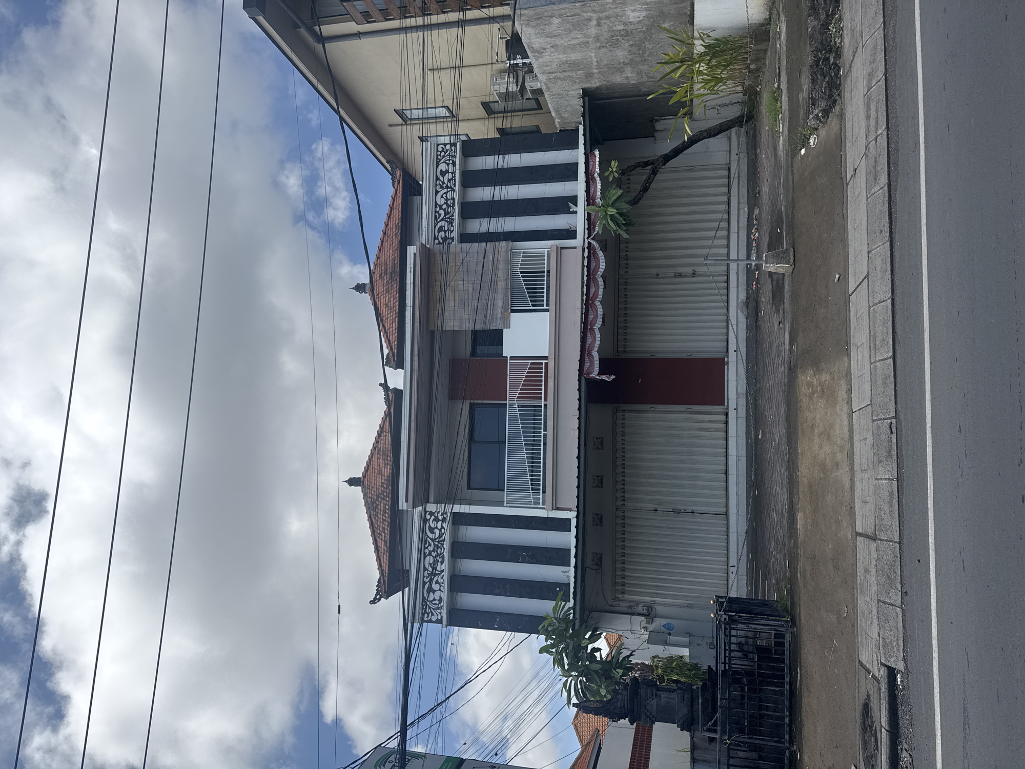 The image depicts a two-story building in North Kuta, Indonesia. The building has a Balinese architectural style, featuring a tiered roof with orange tiles and decorative carvings. The facade is painted with black and white stripes and has a balcony with white railings on the second floor. The ground floor has a closed roller shutter door. An adjacent building is visible on the left, and power lines run across the sky above. The sky is partly cloudy. A narrow sidewalk and a road are visible on the right side of the image. A small garden with plants is present in front of the building. A metal cage is placed close to the garden.