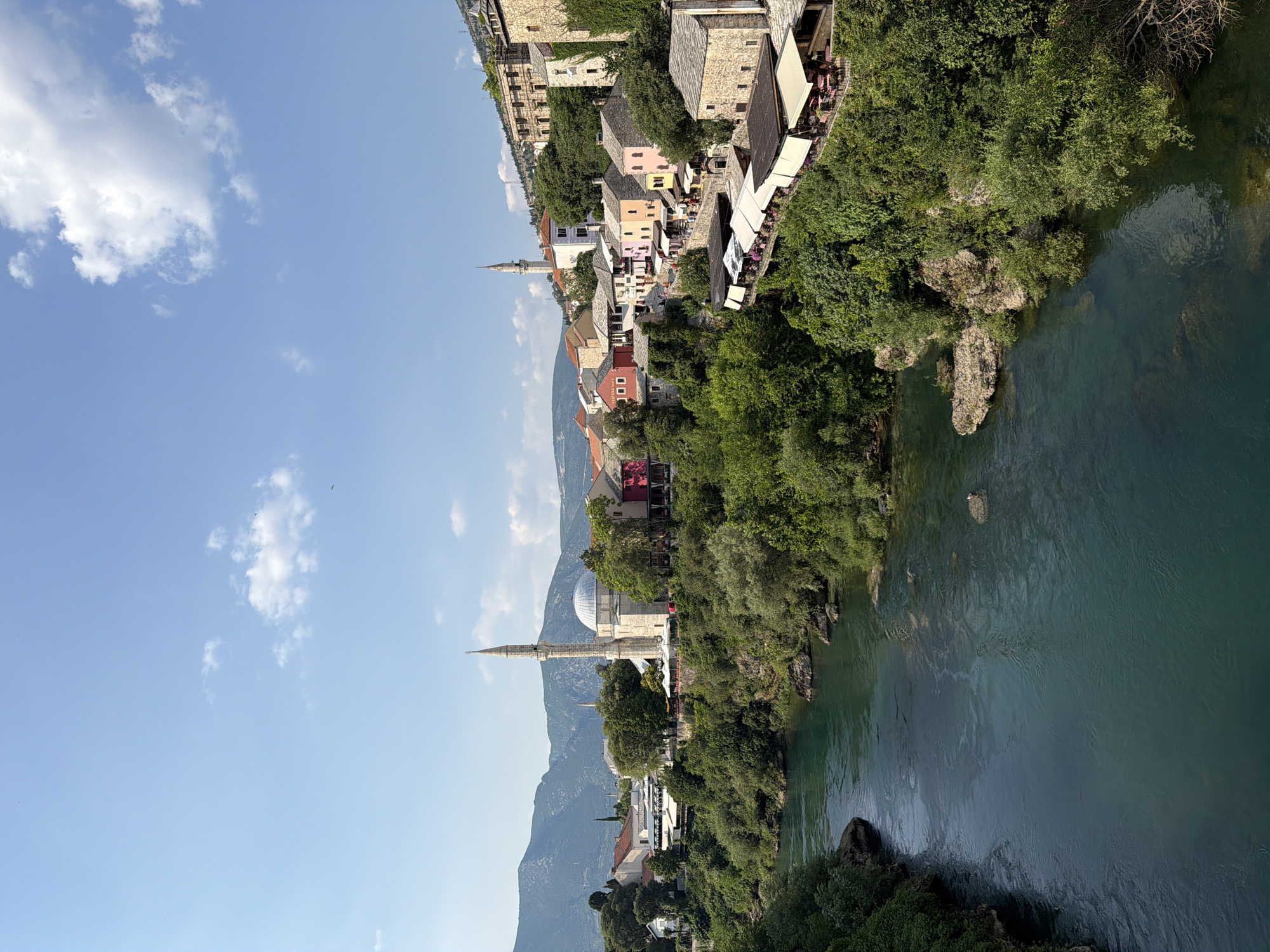 This media file captures a scenic view of Mostar, Bosnia and Herzegovina, with the Neretva River flowing through a gorge. The scene is dominated by a collection of traditional stone buildings lining the riverbank, interspersed with lush green trees. In the distance, a prominent mosque with a minaret and dome stands as a key architectural feature, indicating a historical and cultural setting. The sky is a clear blue with scattered white clouds, suggesting fair weather and a daytime setting, likely mid-morning or afternoon given the sunlight. Rocky outcrops are visible along the river's edge, and the water itself is a deep, tranquil green. The buildings suggest a vibrant community, with outdoor seating areas and awnings indicating active cafes or restaurants, implying a bustling atmosphere. The overall impression is one of picturesque natural beauty combined with rich historical architecture.